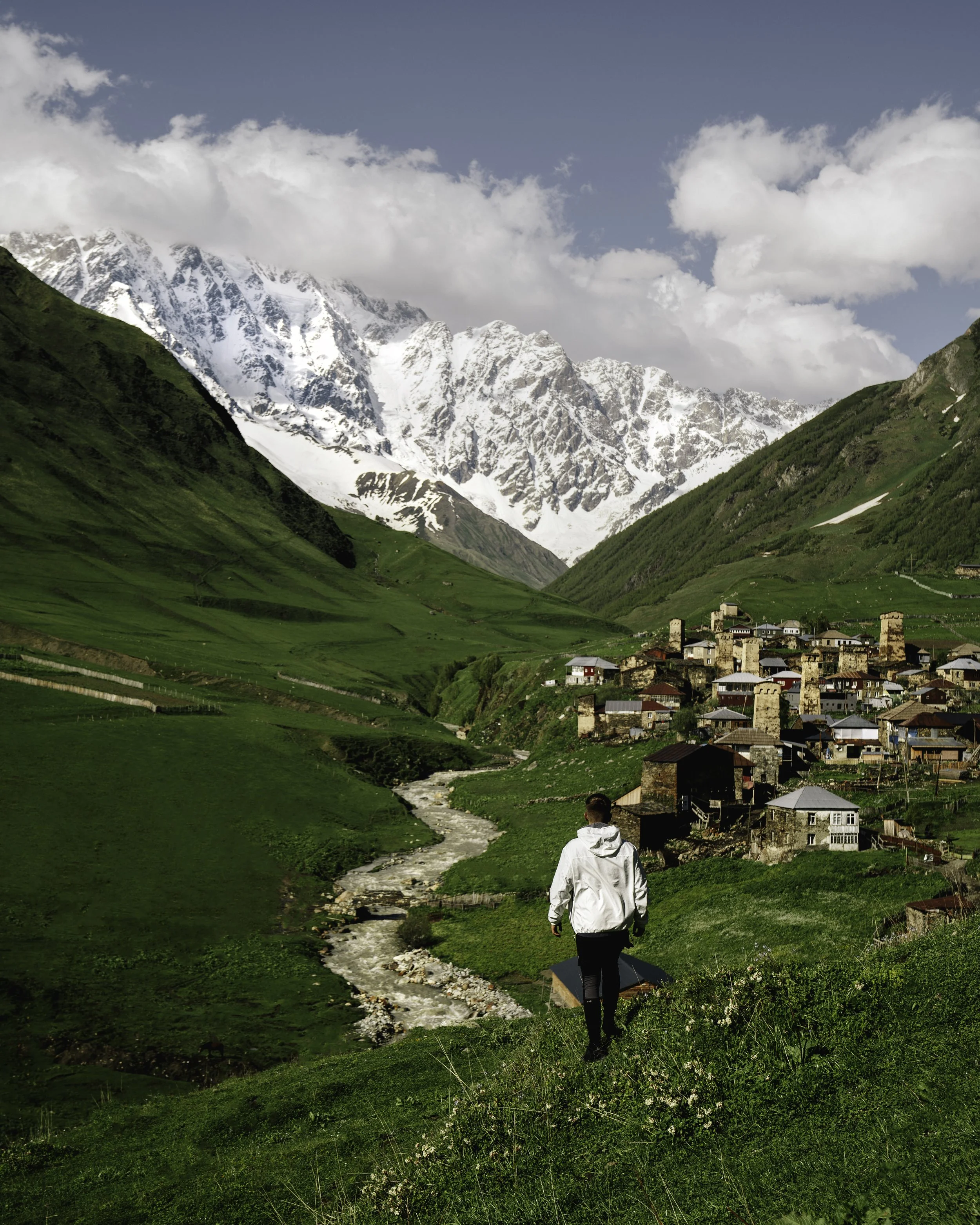 A person walking along a narrow trail in a green valley with a small creek at the bottom, mountain village with stone buildings, and snow-capped peaks in the background under a partly cloudy sky.