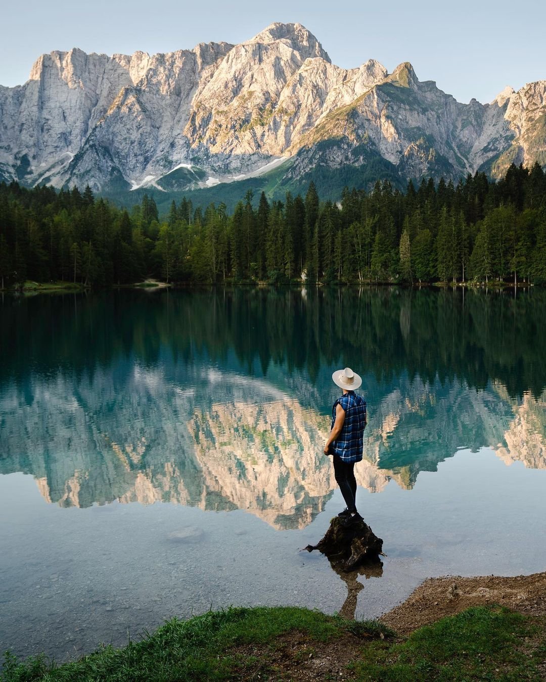 Person standing on a small tree stump near a calm lake, facing mountains and a dense forest in the background, with their reflection visible in the water.