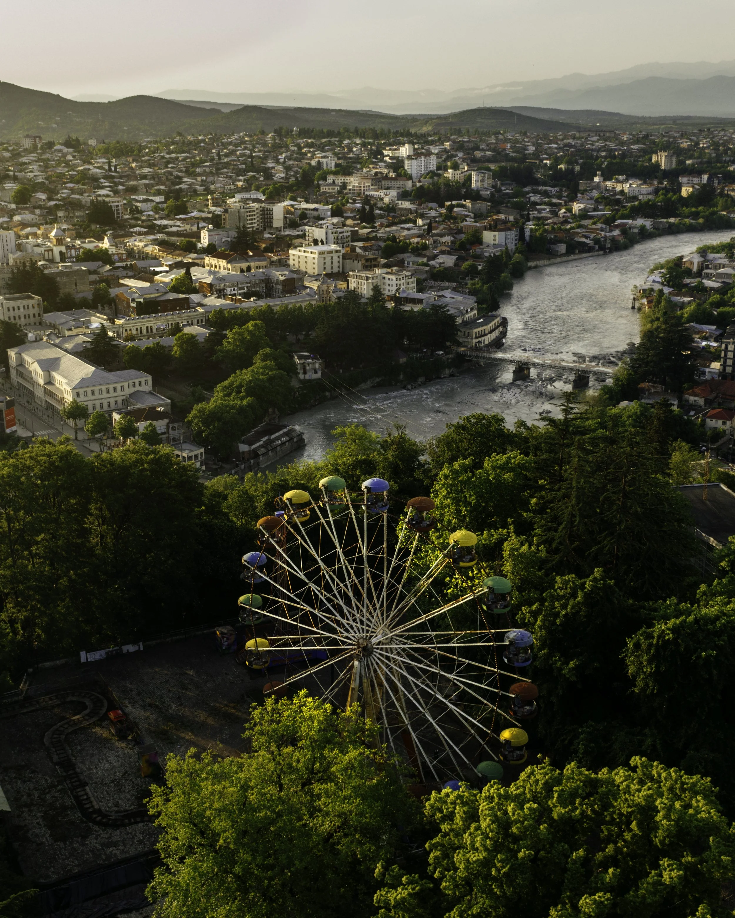 An aerial view of a city with numerous buildings, a river running through it, and a large Ferris wheel surrounded by green trees.