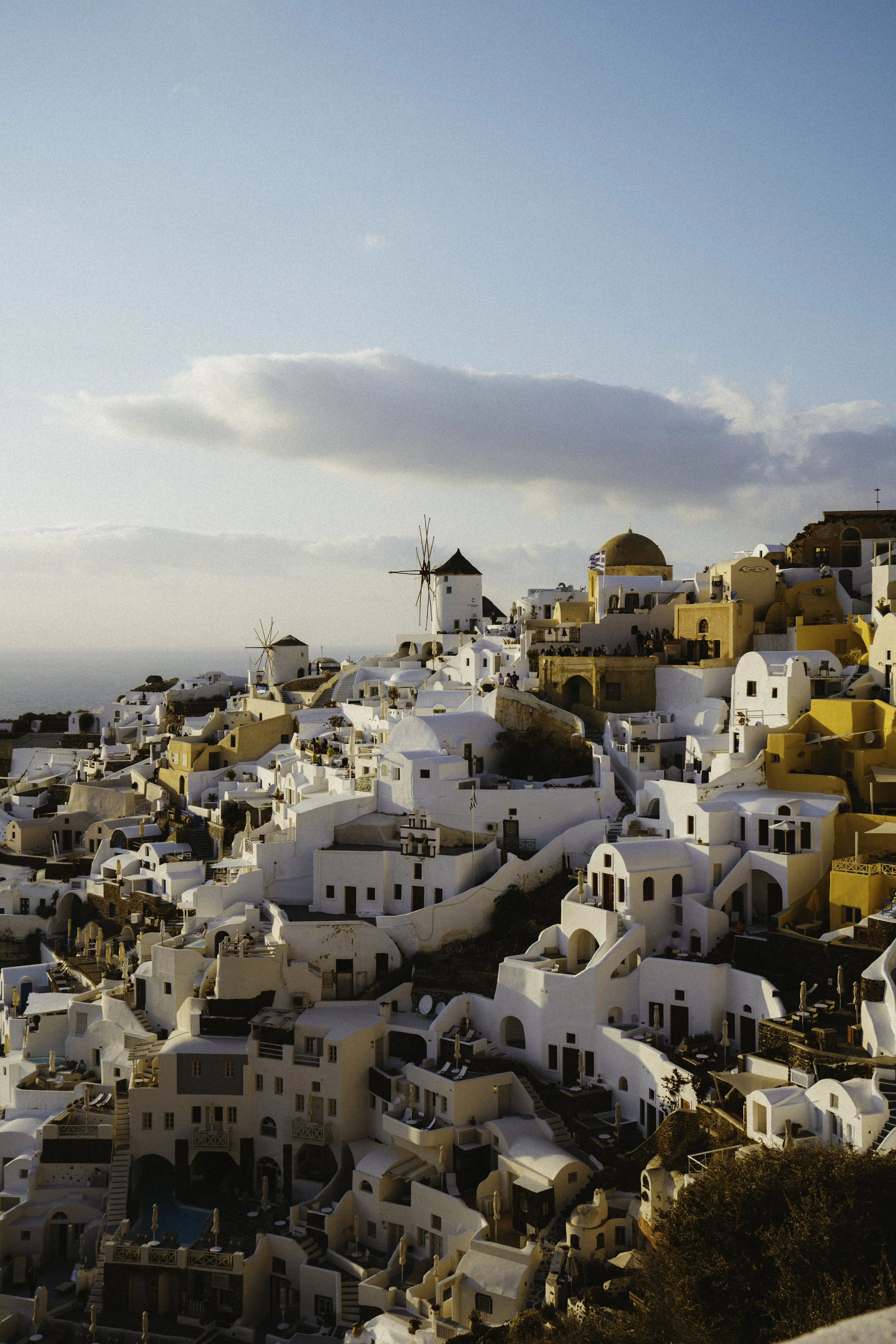 White buildings with blue accents and windmills on a hillside in Santorini, Greece, under a blue sky with clouds.