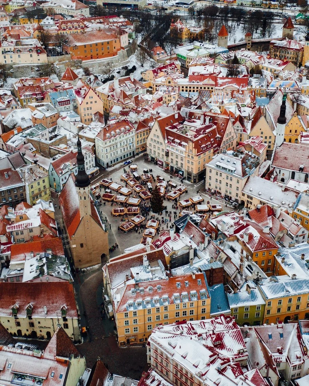 Aerial view of a historic European town square covered in snow, featuring colorful multi-story buildings, church steeples, and market stalls around a decorated Christmas tree.