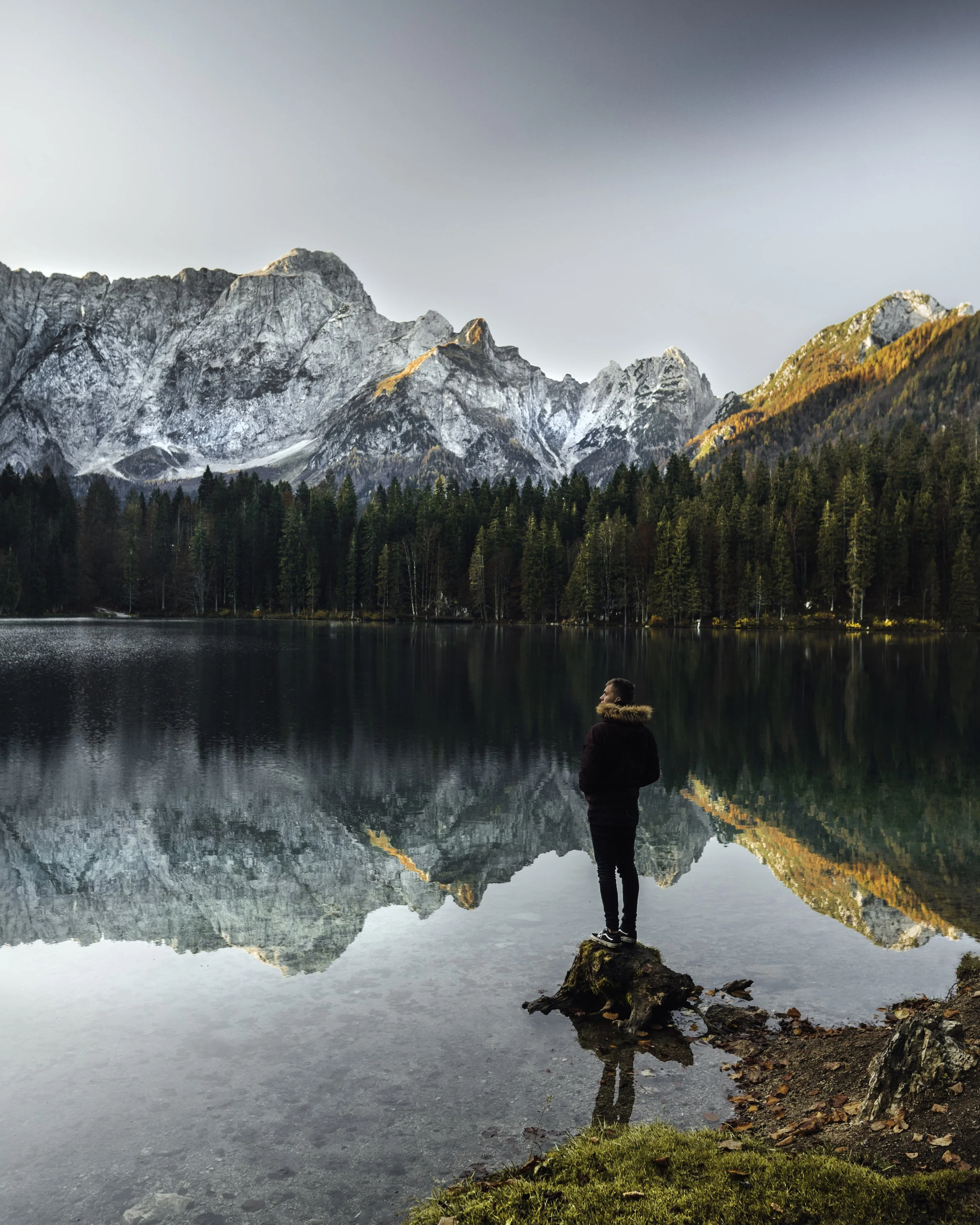 A man stands on a rock at the edge of a lake, with a forest and mountain range in the background, and the mountain's reflection in the water during sunset.