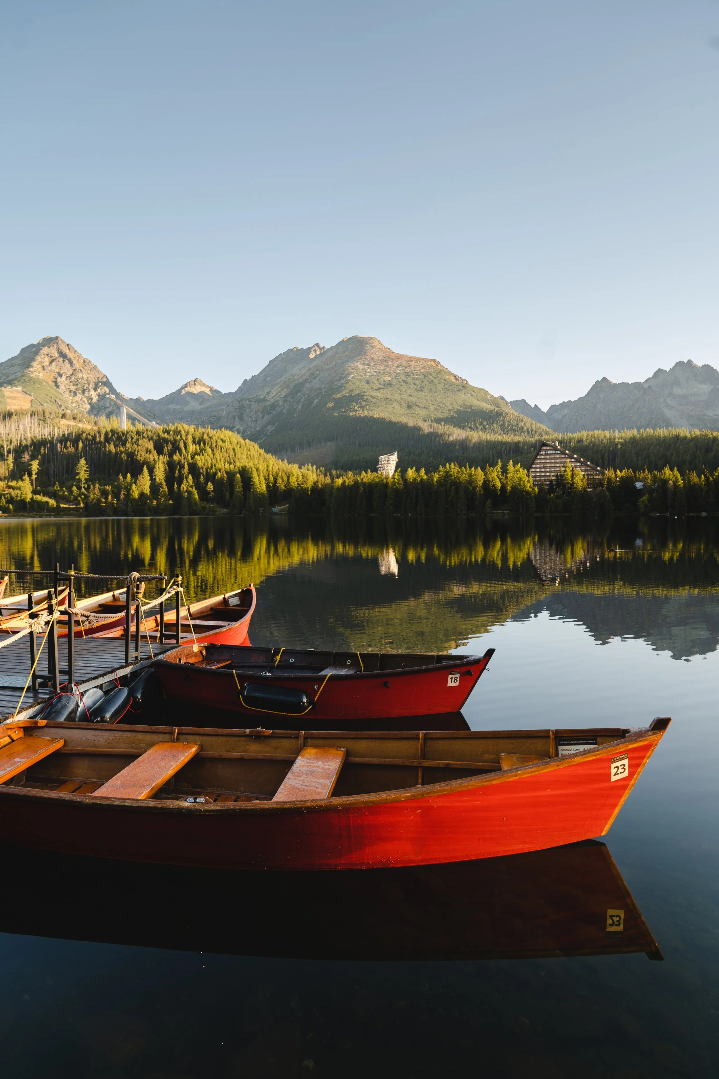 Three wooden and red rowing boats are docked on a calm lake with a backdrop of green forested mountains and clear blue sky.