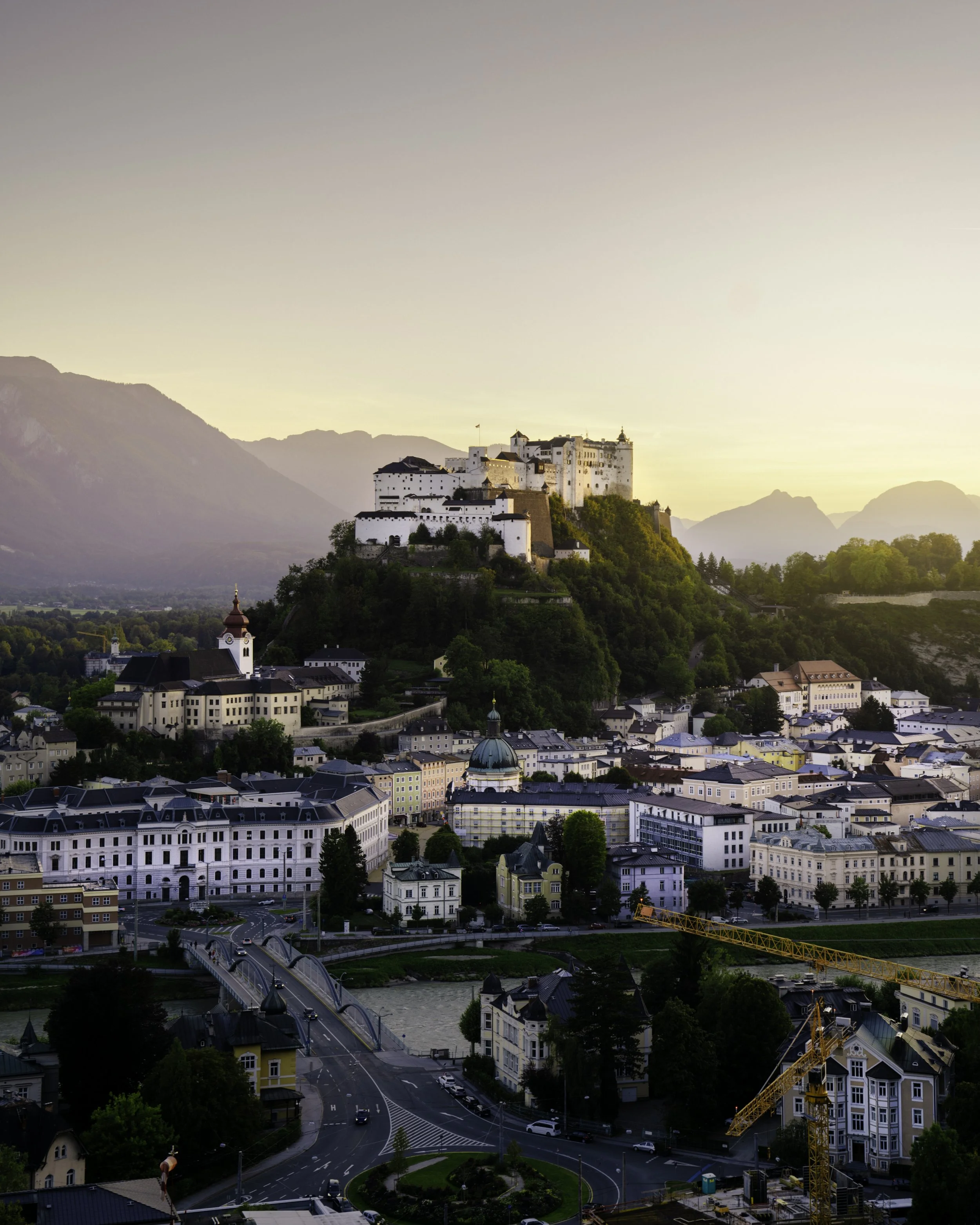 A scenic view of a European city, featuring a hilltop castle and a bridge over a river at dusk, with mountains in the background.