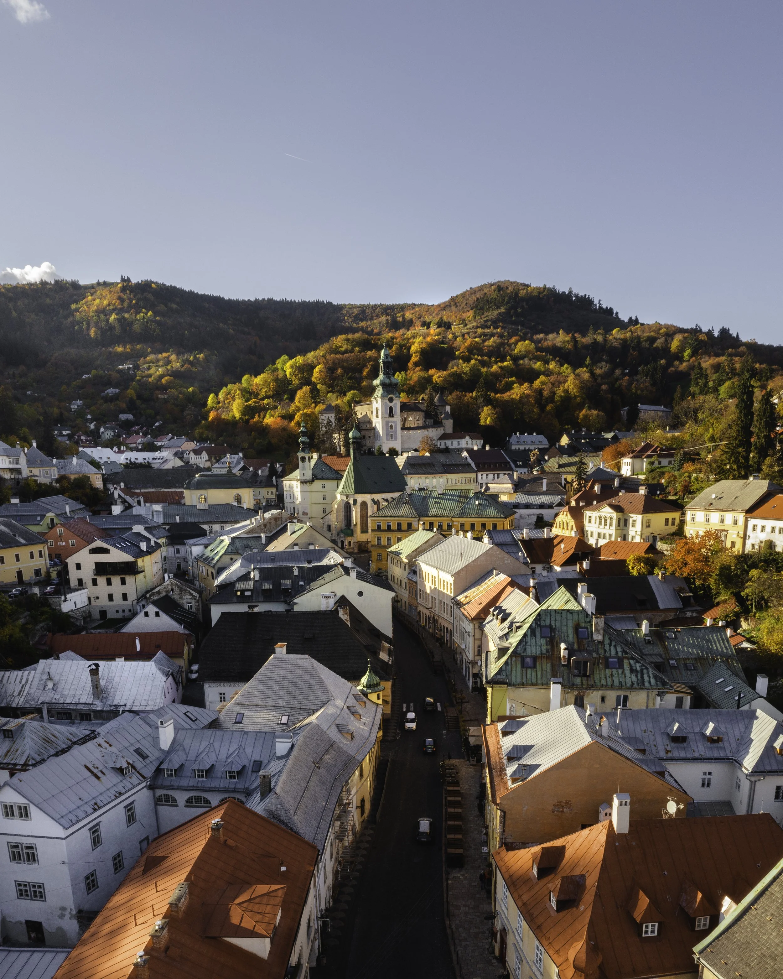 A scenic view of a European town nestled at the foot of rolling hills covered with trees in fall colors, with church steeples and historic buildings in the foreground.