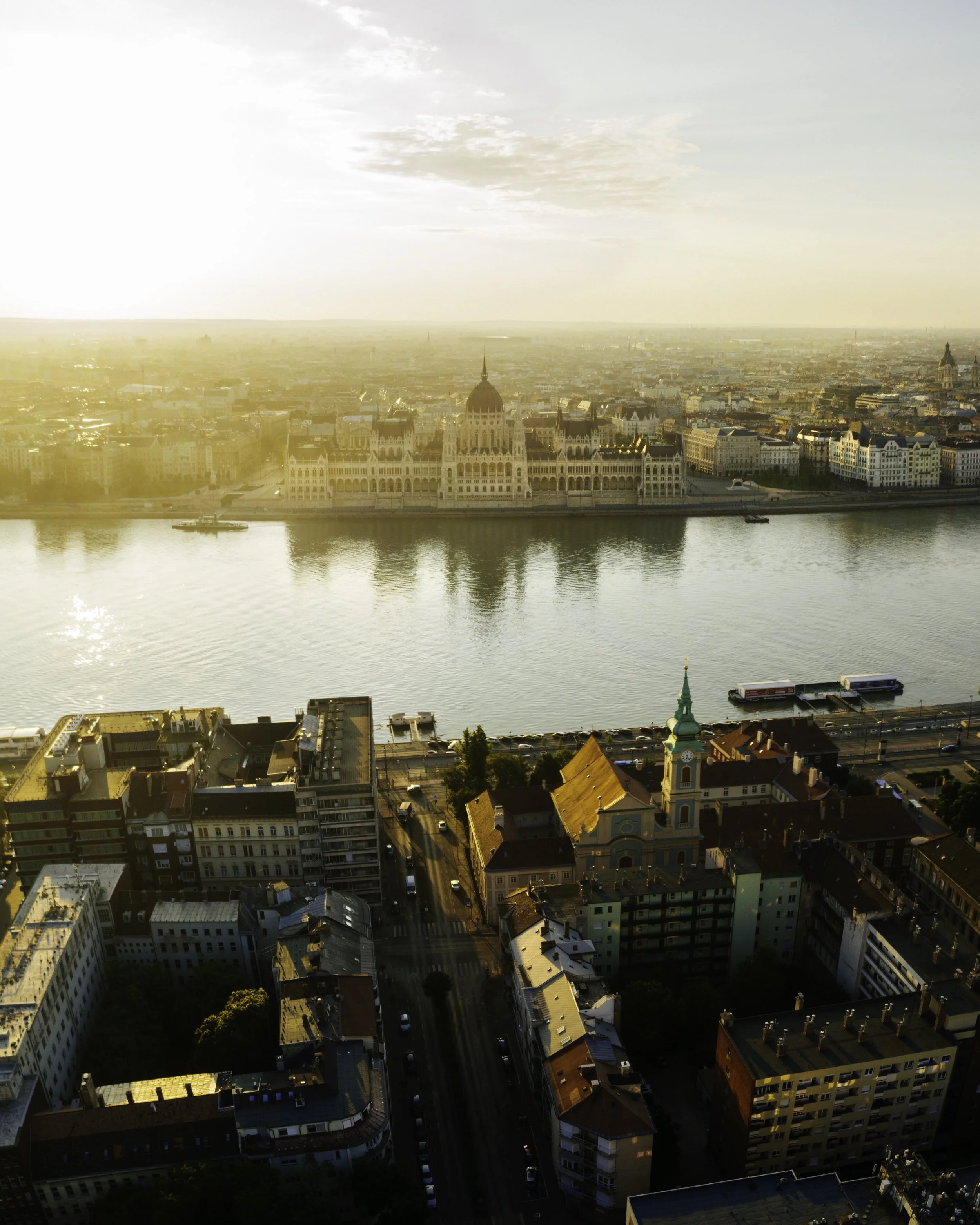 An aerial view of a cityscape at sunset featuring a large historic building along a river, with surrounding modern and historic buildings, and a boat on the river.