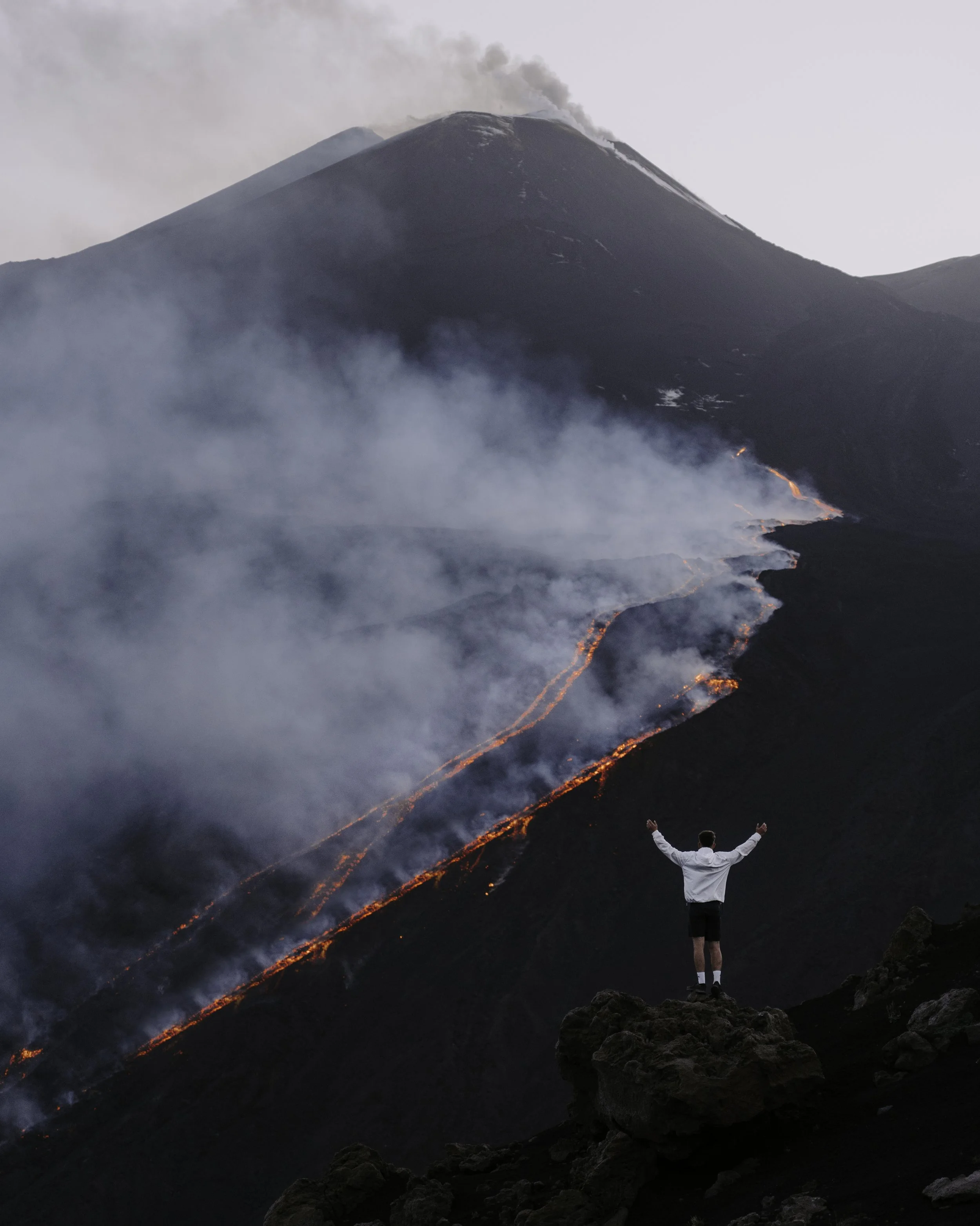 Person standing on rocks with arms raised in front of erupting volcano with flowing lava and smoke.