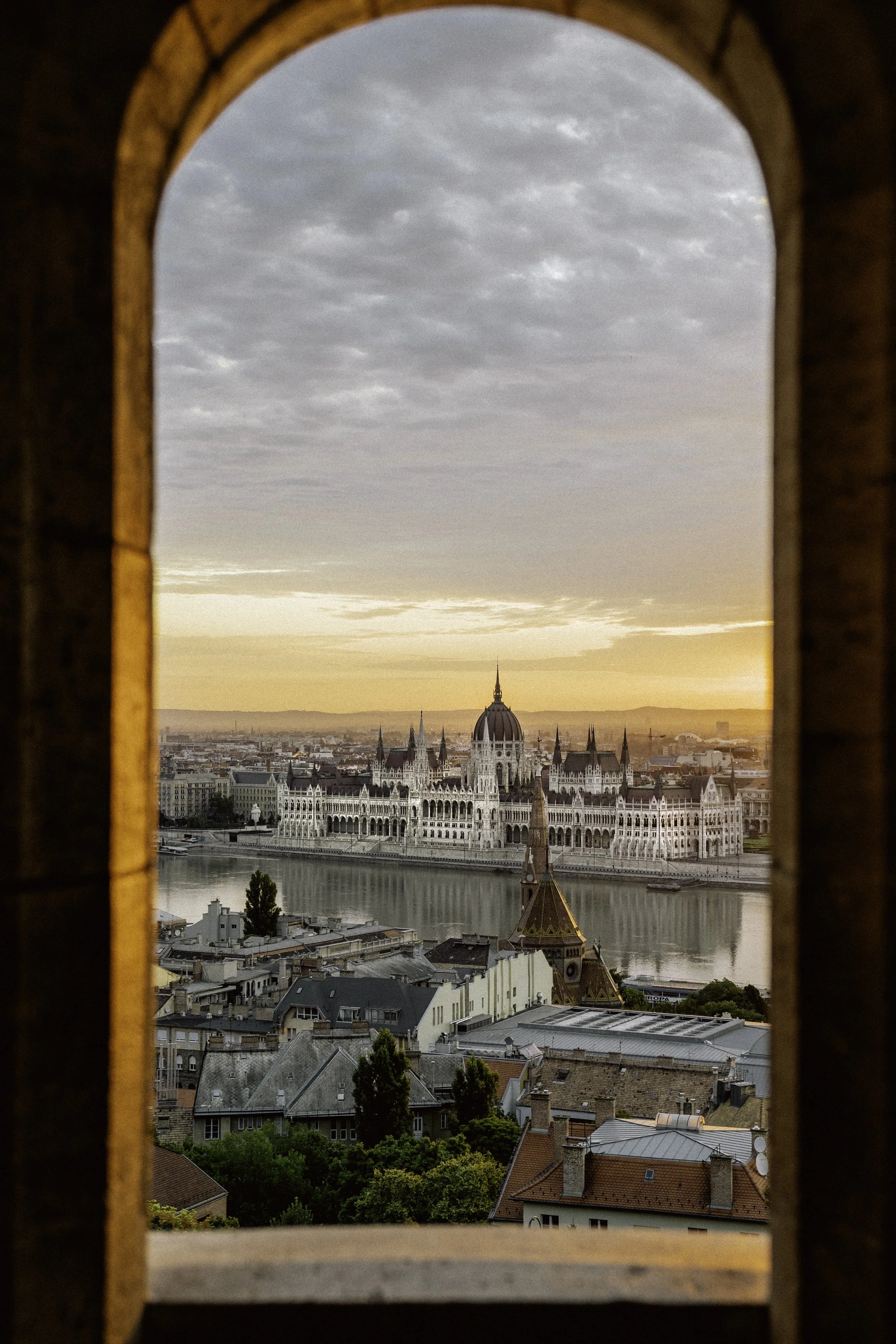 View of the Hungarian Parliament Building at sunrise, seen through a brick arch window, with water in the foreground and a cloudy sky.
