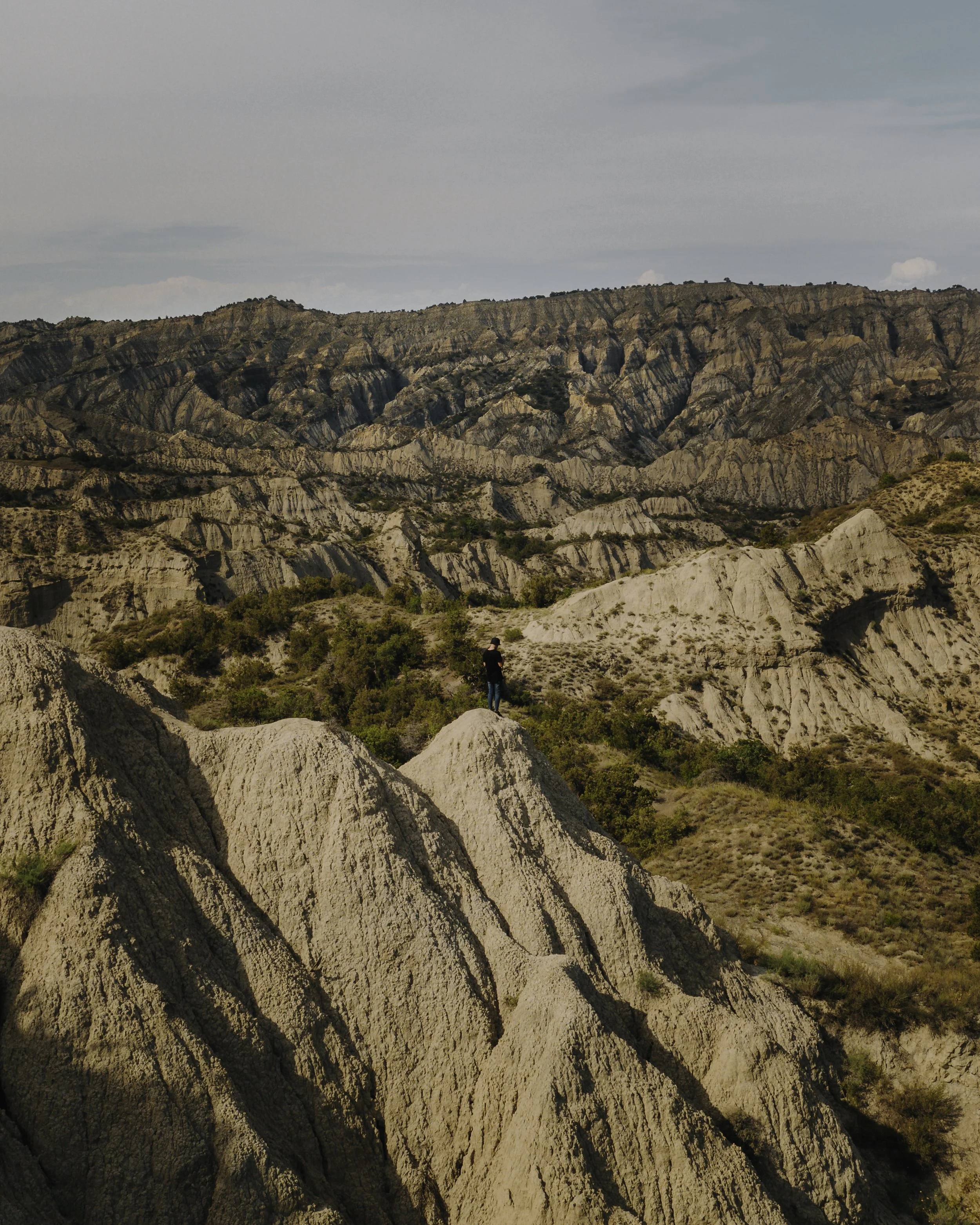A person stands on a rocky hillside overlooking rugged, eroded mountains with layered sedimentary formations, sparse green vegetation, and a cloudy sky above.