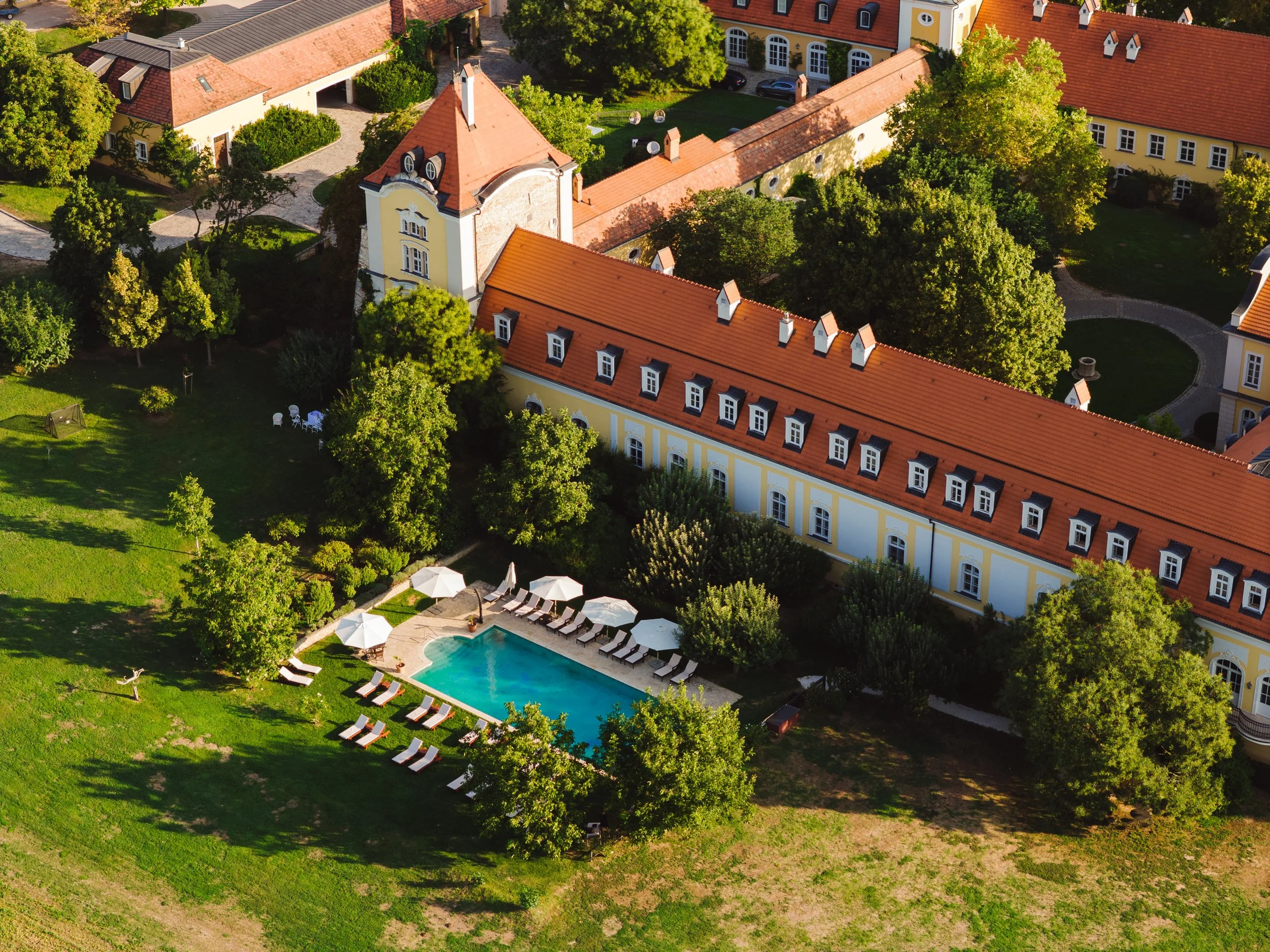 An aerial view of a large, elegant estate with a yellow building featuring red tile roofs, a swimming pool surrounded by lounge chairs and umbrellas, lush green trees, and manicured lawns.