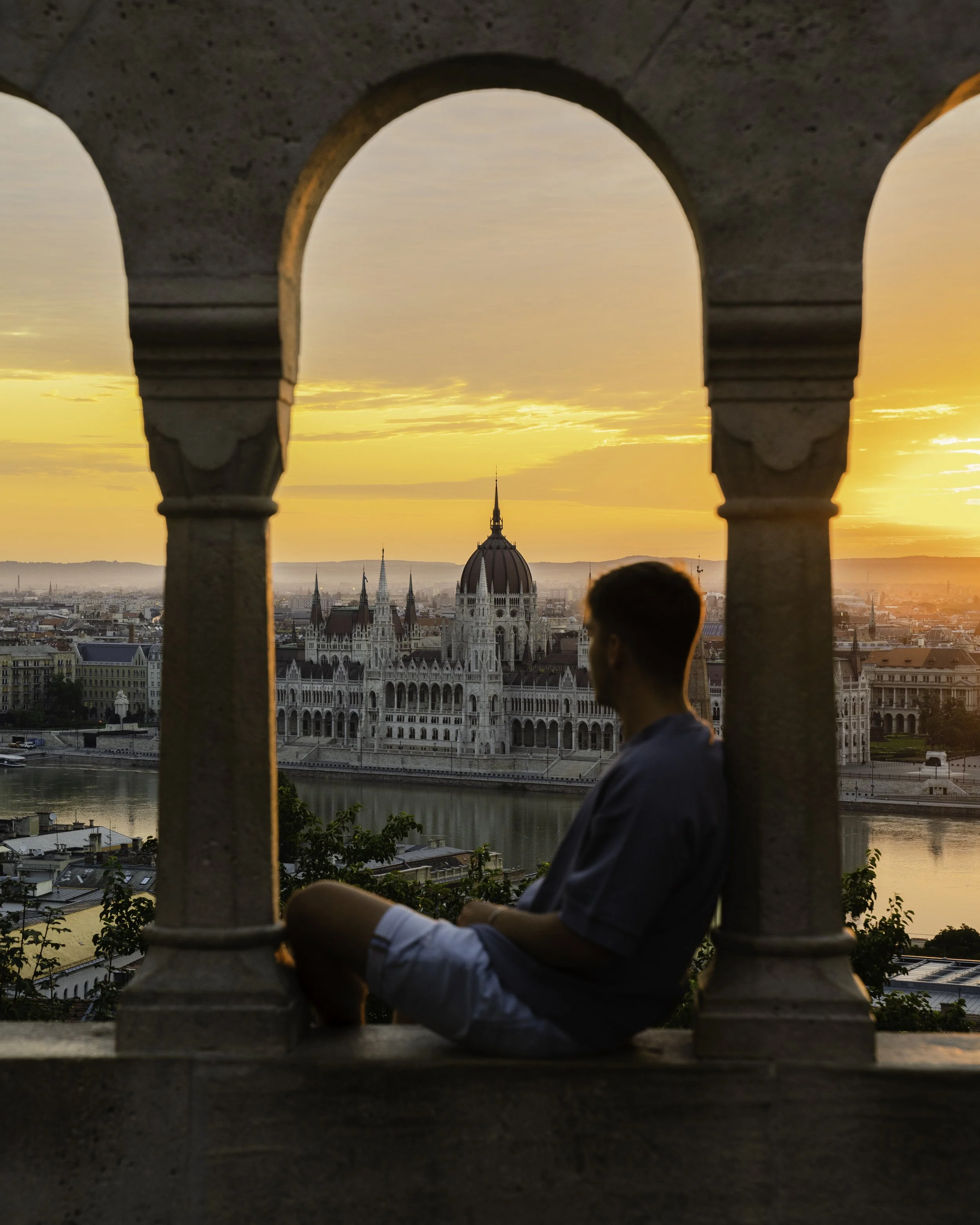 A man sitting on a ledge under a stone archway at sunset, overlooking a cityscape with historical buildings and the Parliament building with a dome in Budapest, Hungary.