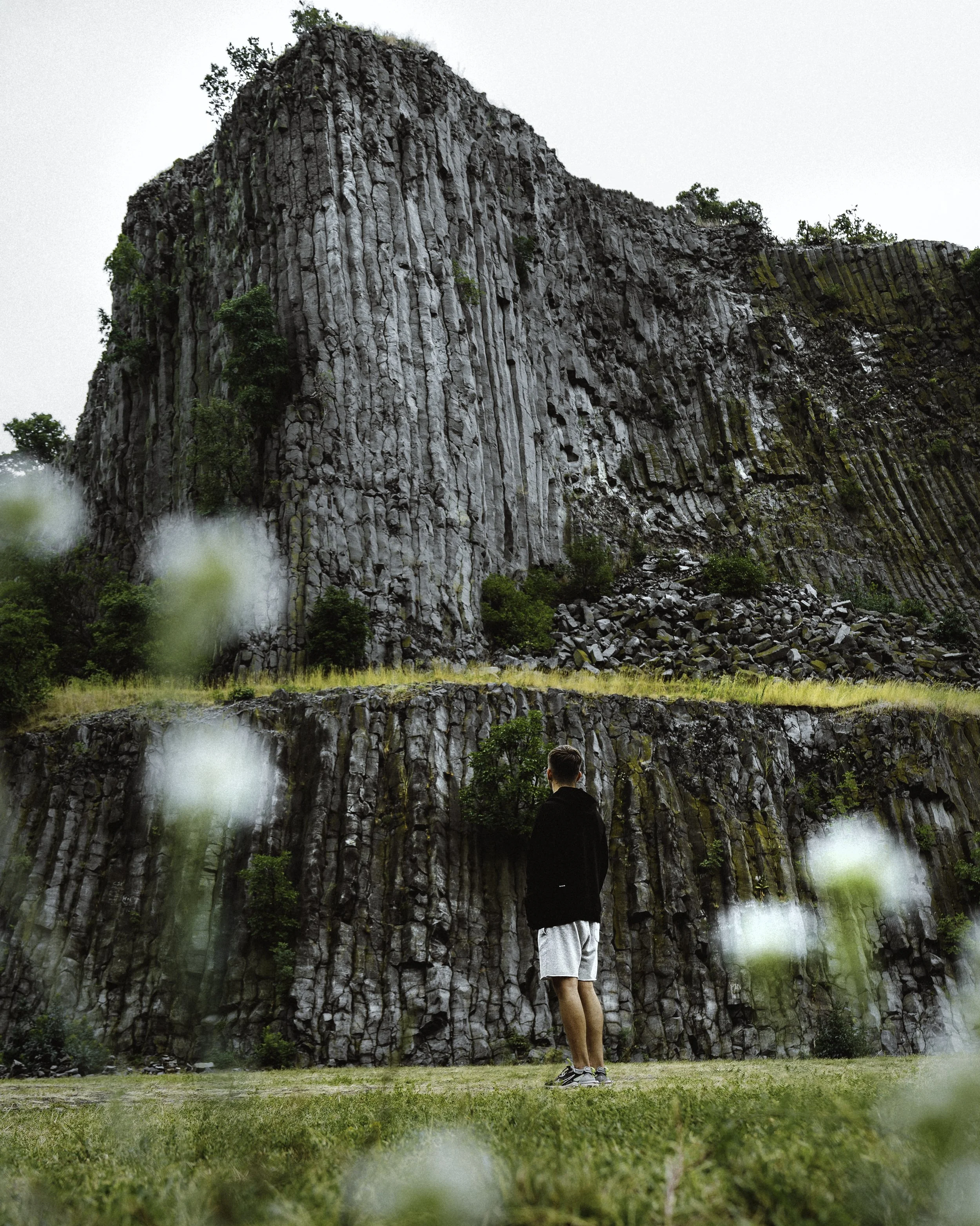 A man wearing a black jacket and white shorts standing on grass in front of a large, textured rock formation with small trees and greenery growing on and around it.