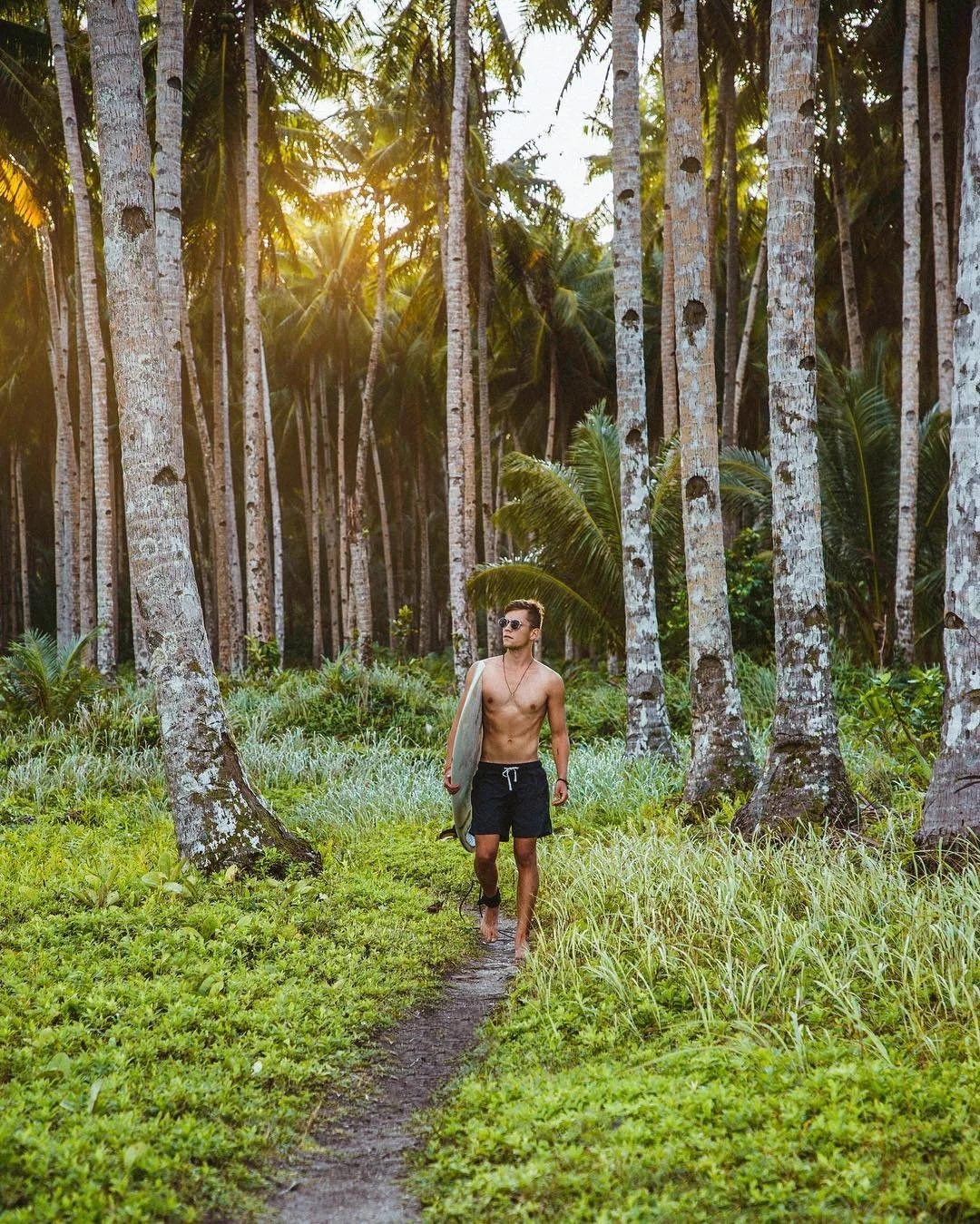 A shirtless man wearing sunglasses and black shorts walking through a tropical forest with tall palm trees and lush green vegetation during sunset.