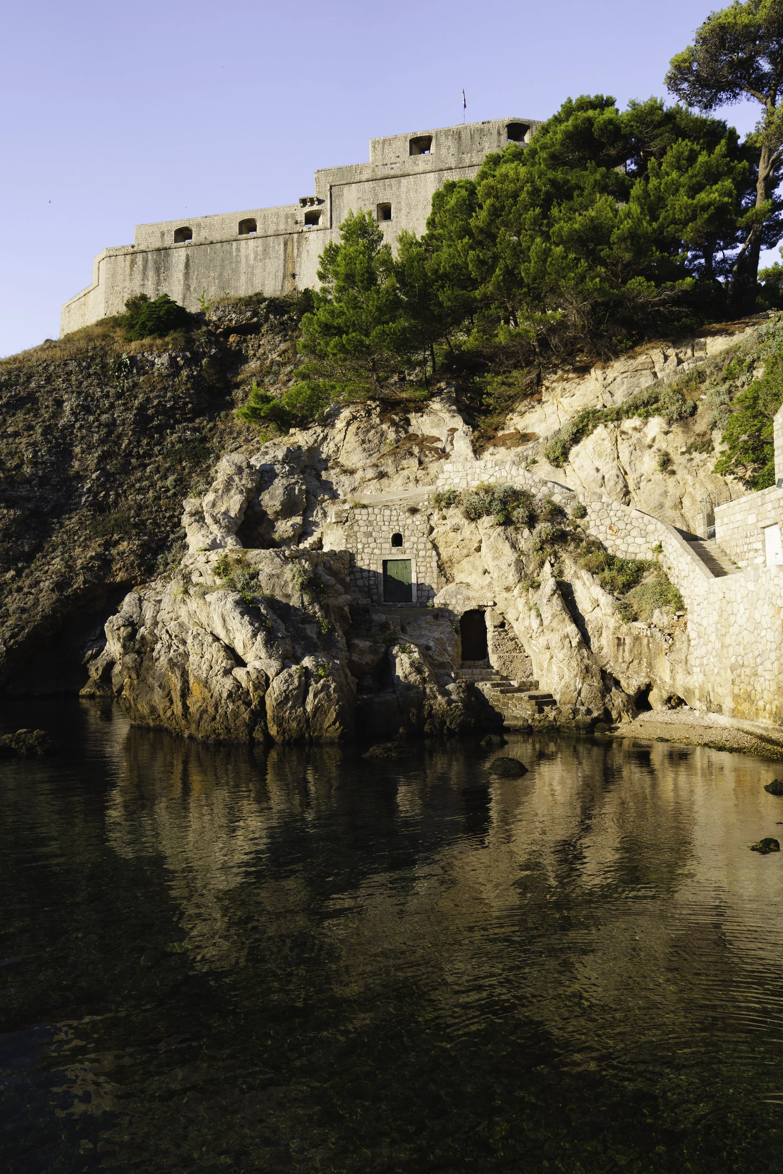A historic stone fortress on a hilltop surrounded by green trees, with steps leading up and water reflecting the fortress at the base during daytime.