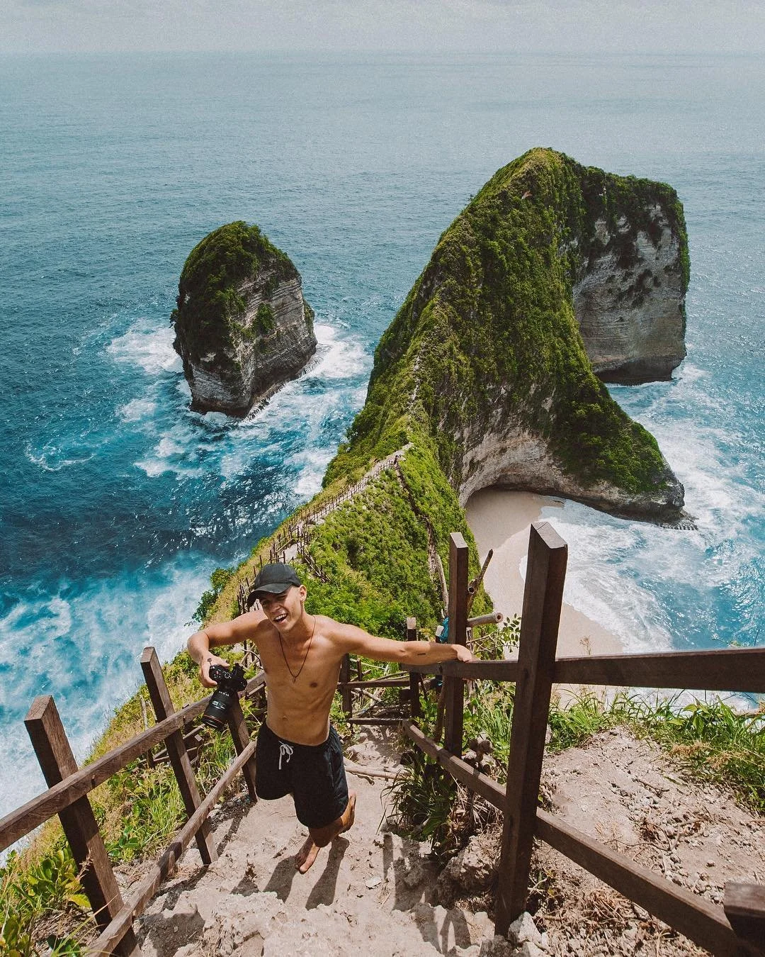 A young man without a shirt, wearing black shorts and a cap, smiling and holding a camera, standing on a staircase overlooking a scenic coastal landscape with limestone cliffs and a beach.