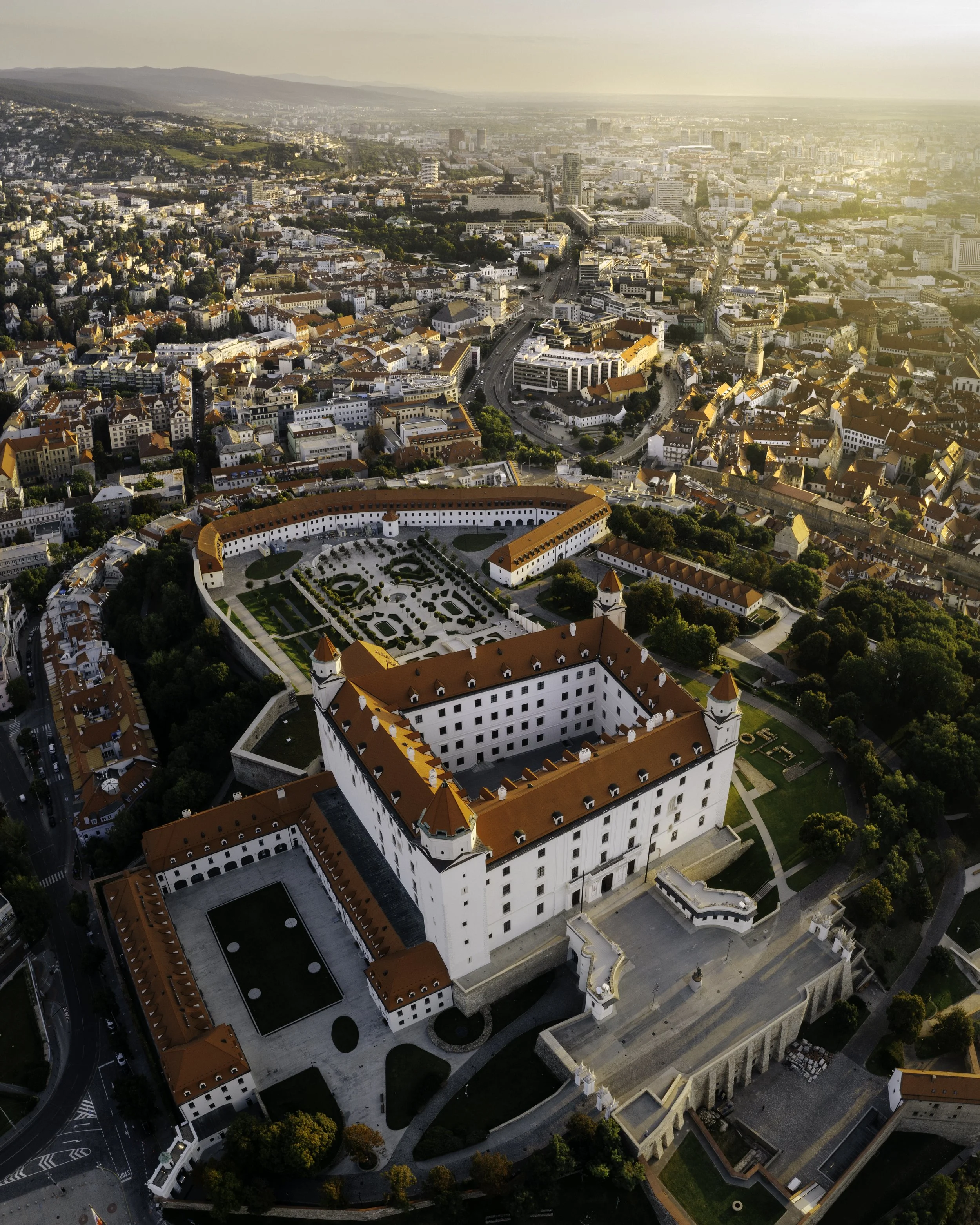 Aerial view of Bratislava Castle in Slovakia with surrounding cityscape and mountains in the background, during sunset.
