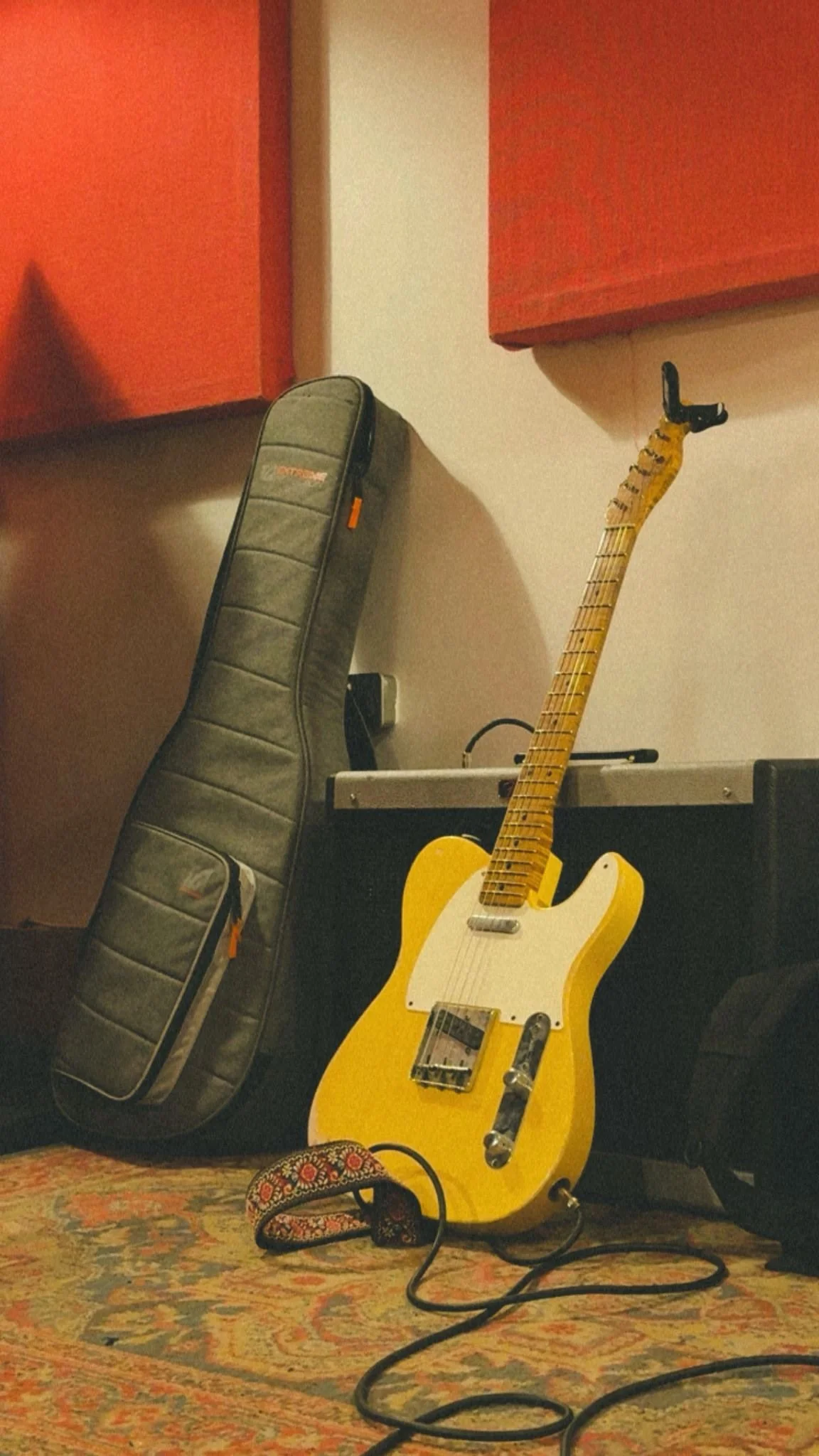 Yellow electric guitar with a white pickguard, resting on a stand, with a guitar strap nearby. A black guitar case leaning against a wall with red acoustic panels, and a patterned rug on the floor.