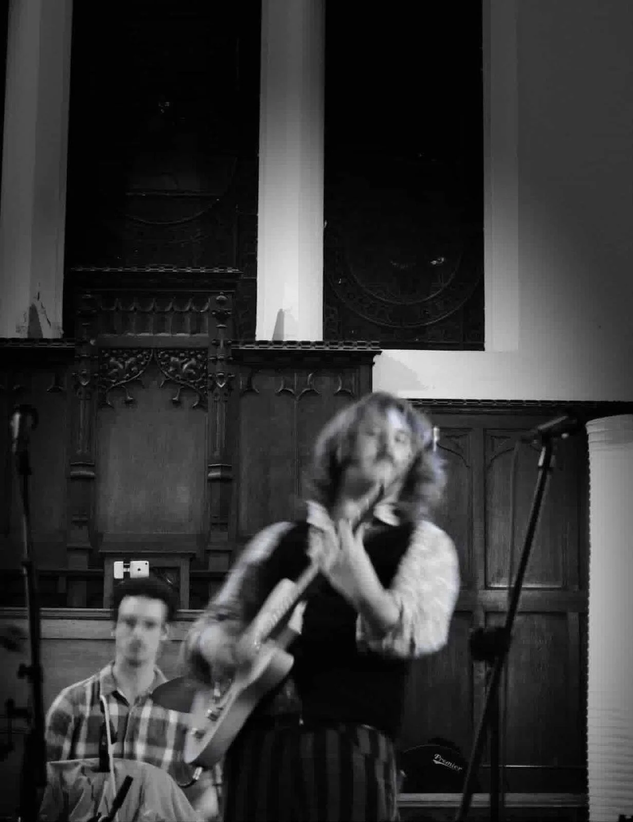 Black and white photo of a woman playing a wind instrument, possibly a flute or clarinet, with a man sitting nearby, in front of a dark wooden wall with decorative carvings, microphones on stands, and a woman in the background.