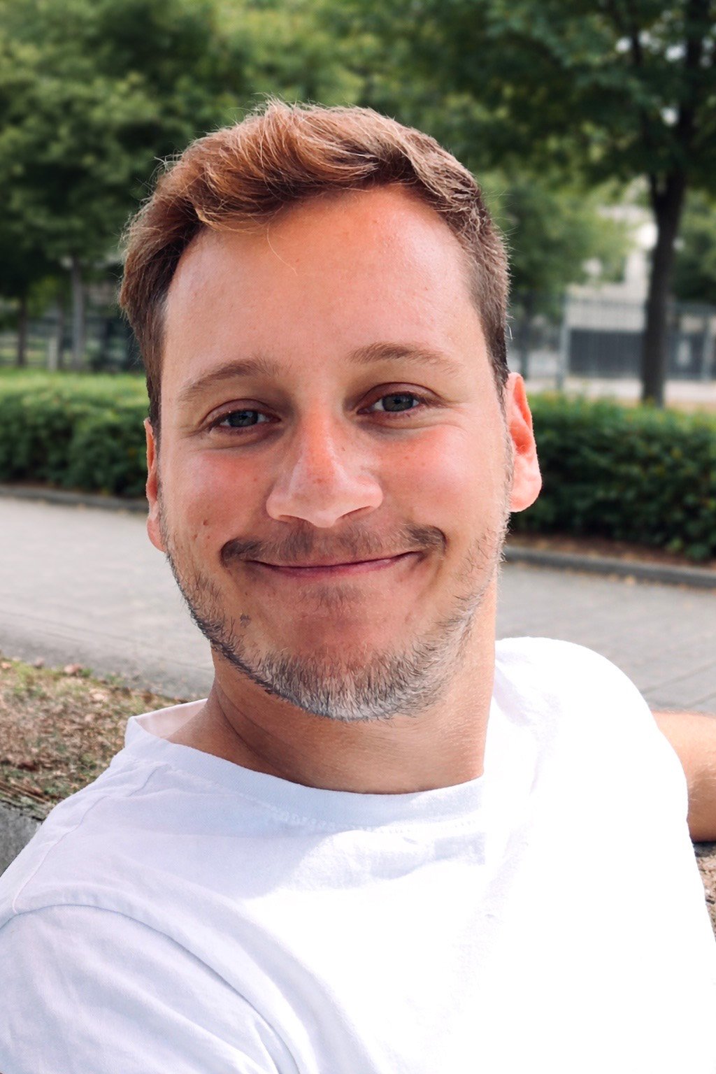 Jeune homme souriant avec des cheveux bruns, portant un t-shirt blanc, dans un parc avec des arbres verts en arrière-plan.