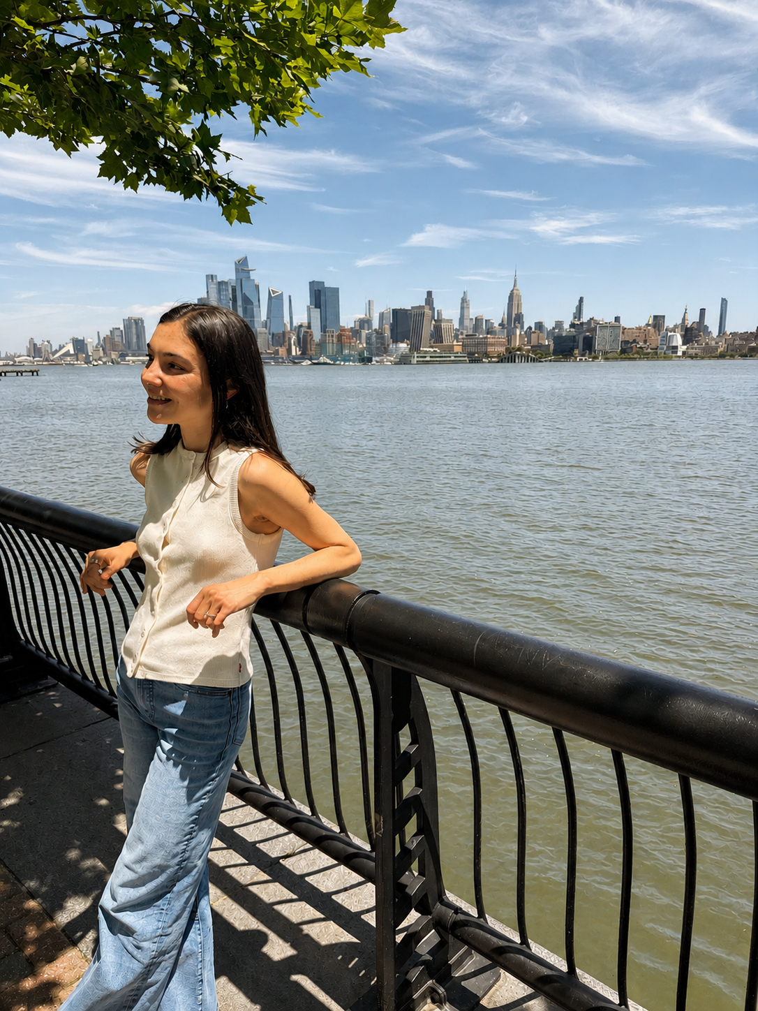 Alexa Nichols, registered dietitian, leaning against a waterfront railing and smiling, with the New York City skyline visible across the river on a sunny day