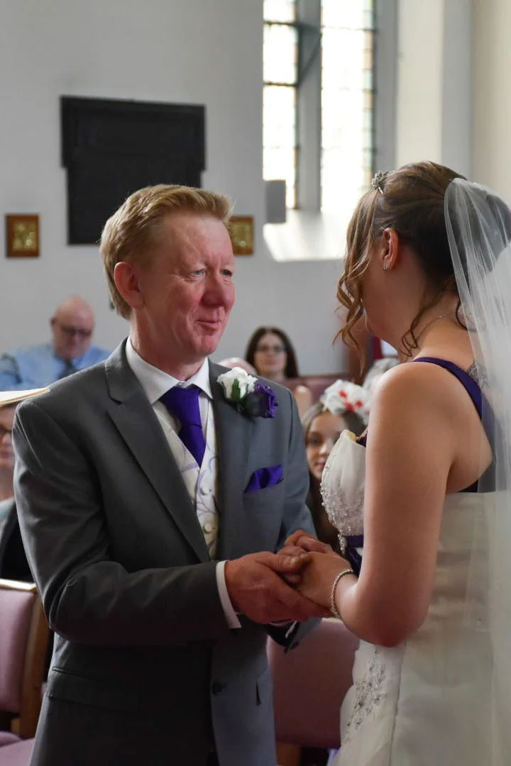 A wedding ceremony with a groom in a gray suit and a bride in a white wedding dress, holding hands and exchanging vows inside a church.
