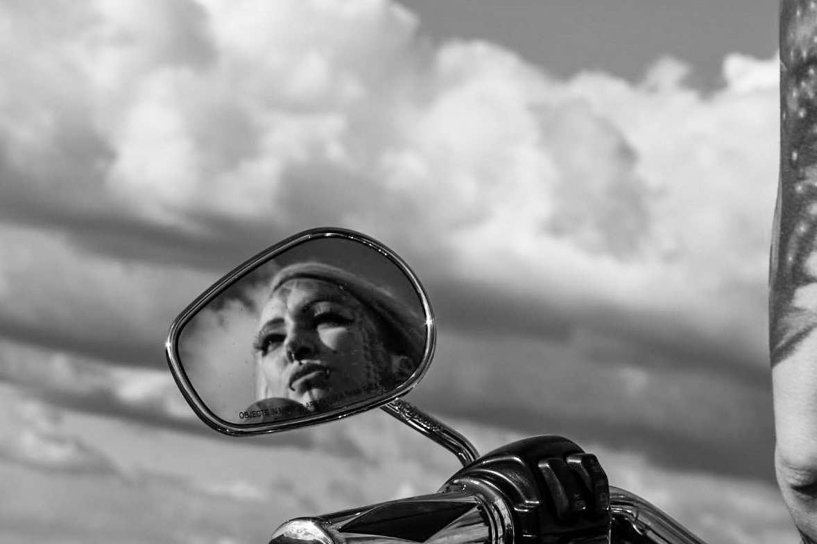 Black and white photo of a mirror on a motorcycle reflecting a woman's face with dramatic makeup and a hat, against a cloudy sky background.