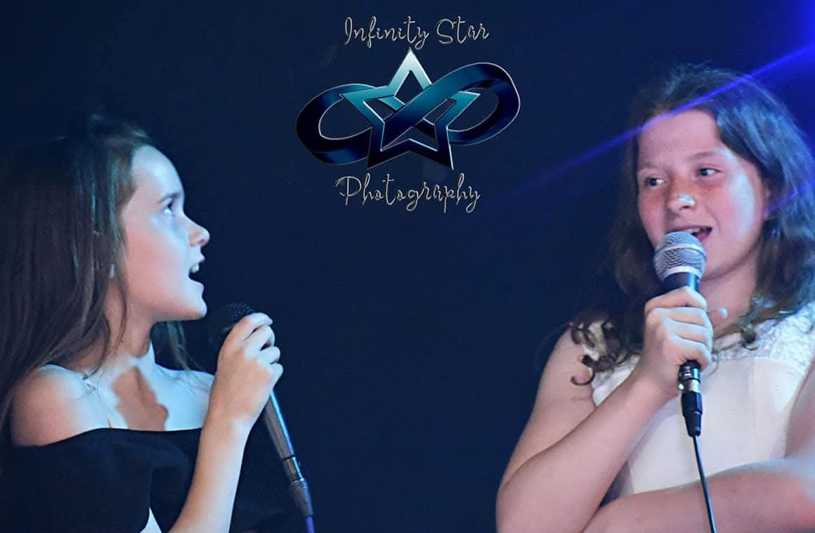 Two young girls are speaking or singing into microphones on stage, with a dark background that has the words "Infinity Star Photography" and a star logo.