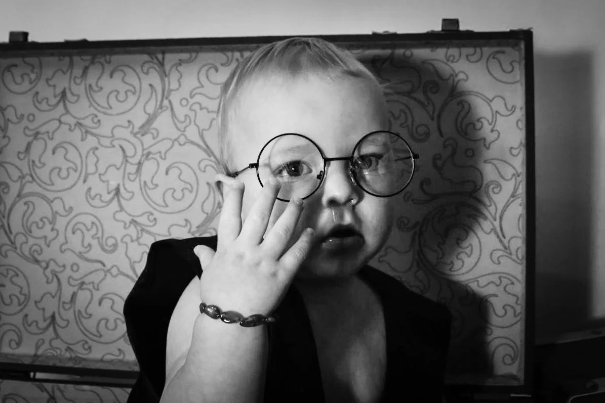 A young child with glasses, touching her face, in front of a decorative patterned backdrop, in black and white.