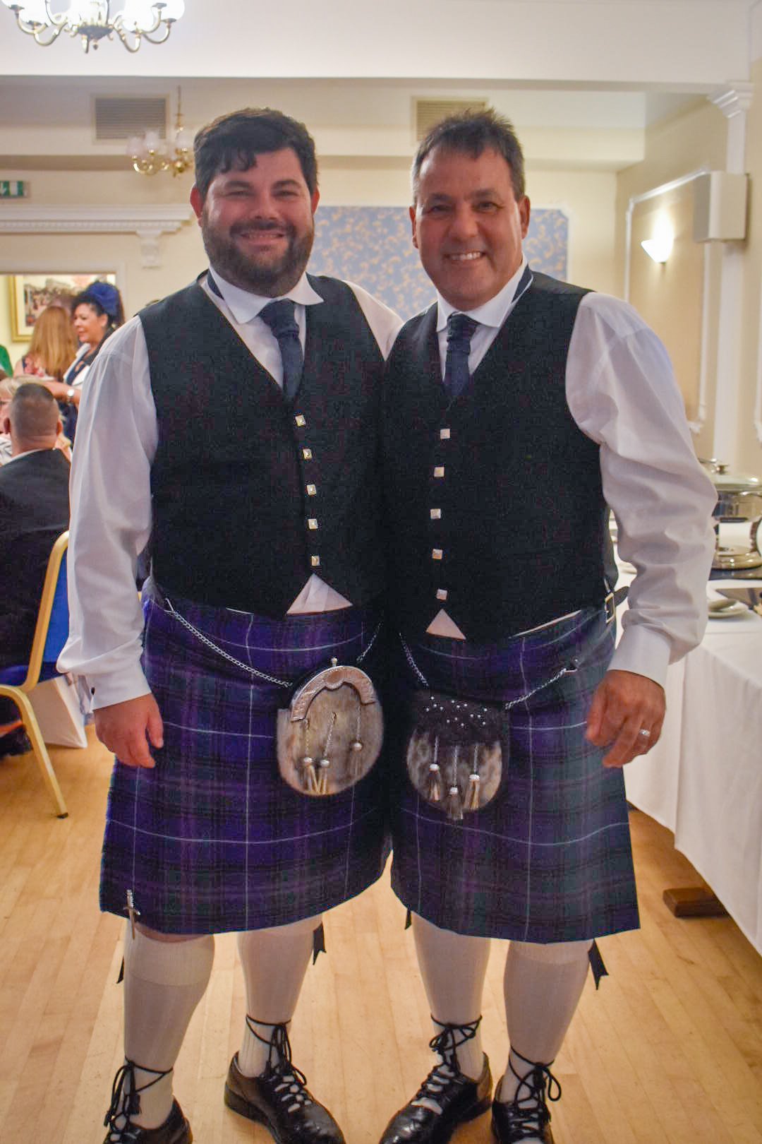 Two men in traditional Scottish kilts and vests, standing indoors at a celebration or gathering, smiling at the camera.