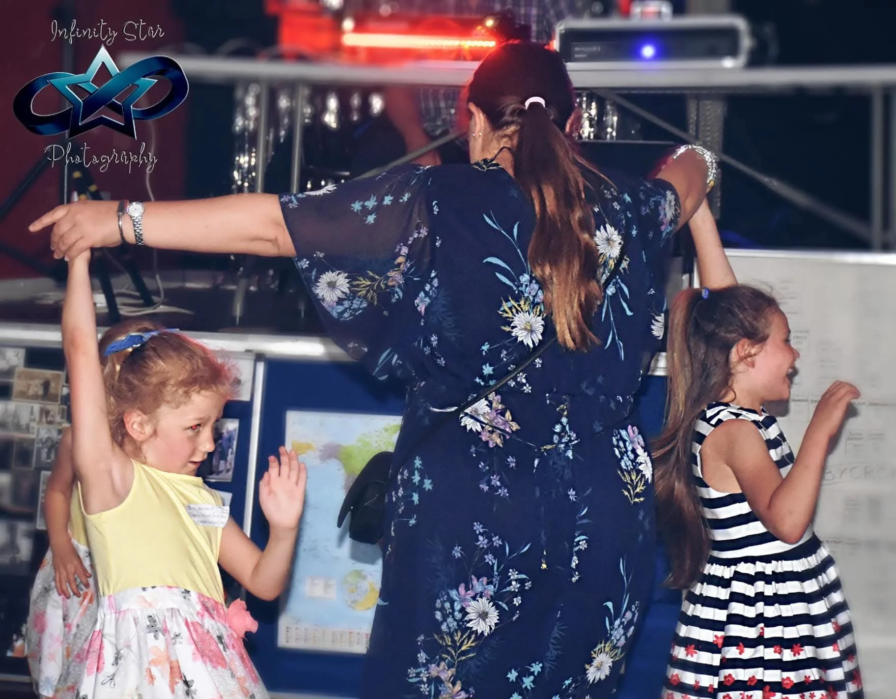 A group of children and an adult are dancing or playing together indoors. The children are smiling and engaging in a lively activity, with a woman leading or guiding them. The background includes a wall with posters and a map, and some electronic equipment is visible. The scene appears joyful and energetic.