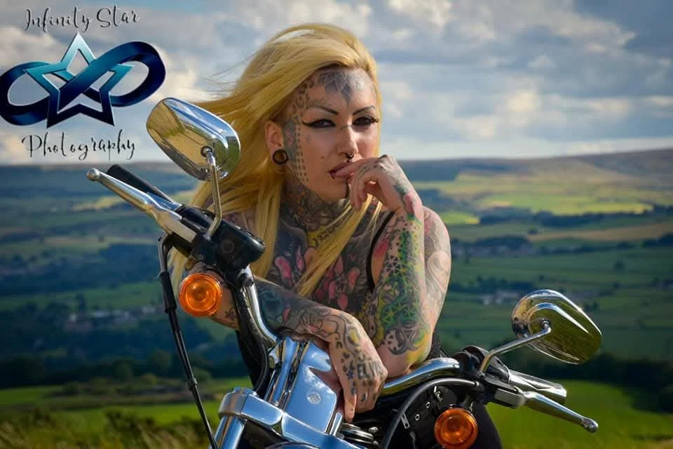 Tattooed woman with long blonde hair posing with a motorcycle outdoors, with rolling green hills and a cloudy sky in the background.