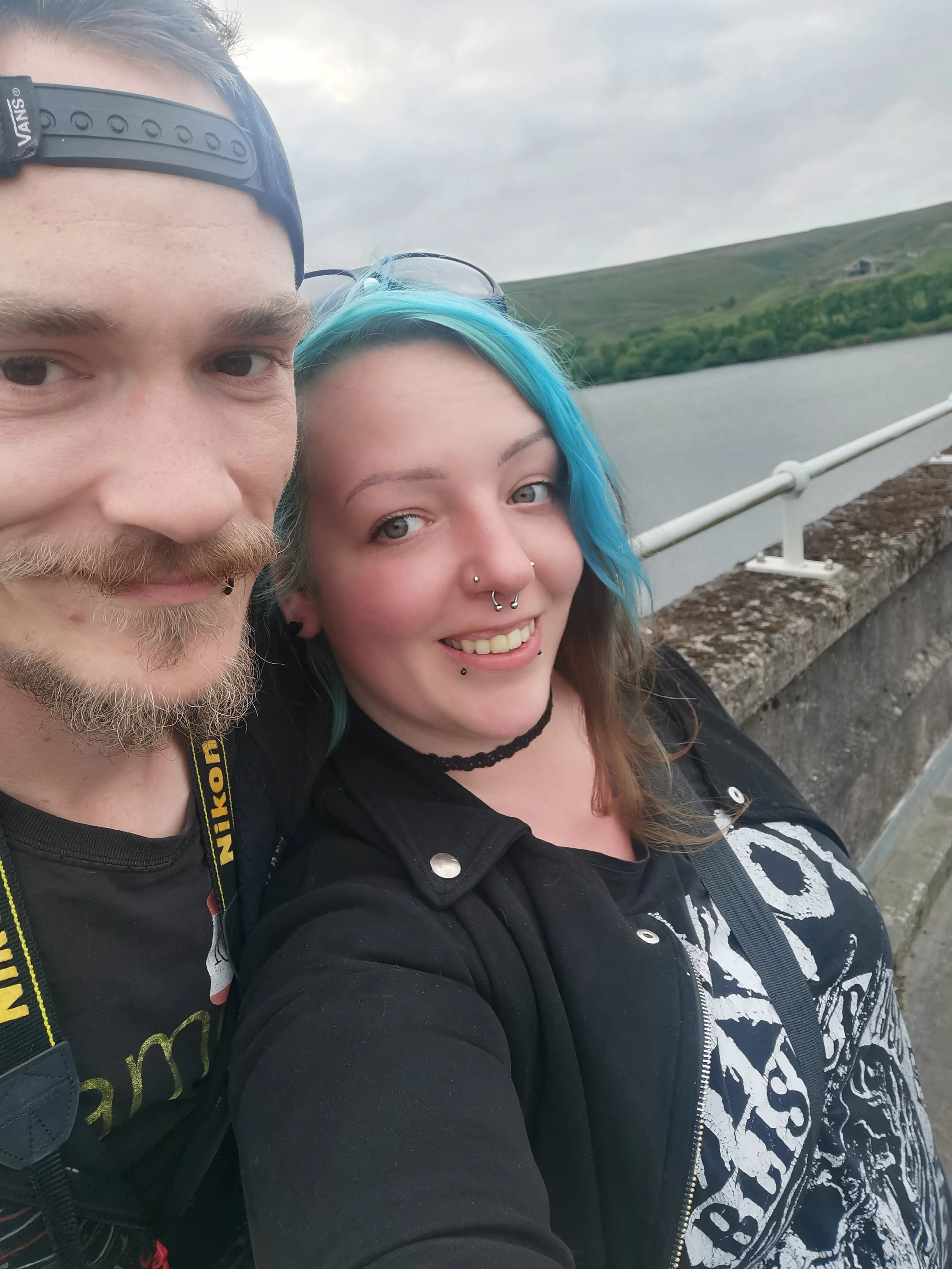 A close-up selfie of a young man and woman outdoors near a body of water with green hills in the background. The man has a beard, light-colored hair, and is wearing a backward baseball cap. The woman has painted blue hair, multiple piercings in her nose and ears, and is wearing a black choker and a black and white patterned zip-up hoodie.