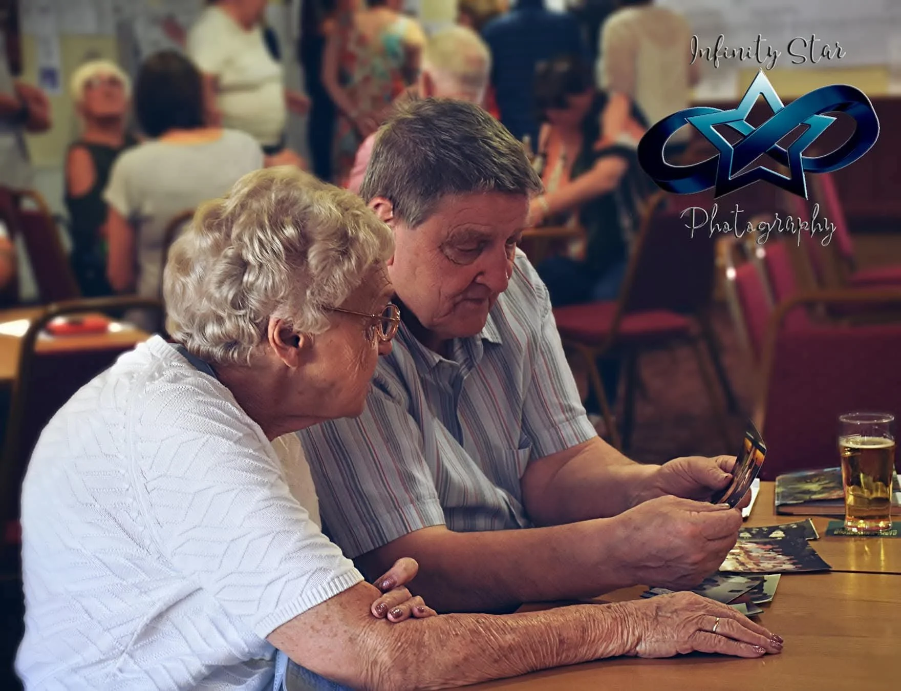 Two elderly people, a woman and a man, sitting at a table looking at a smartphone together in a social gathering or restaurant setting, with other people in the background.