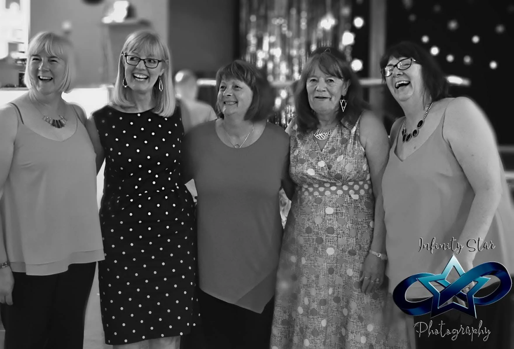 A group of five women standing together smiling, dressed in semi-formal attire, in an indoor setting with holiday decorations and a background of blurred lights.