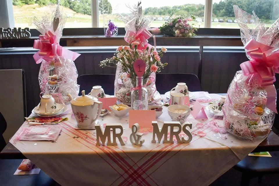 A decorated wedding or reception table with floral arrangements, tea cups, a teapot, and a sign reading "MR & MRS". Pink ribbons and wrapped gift bags decorate the table, which is set near a window overlooking a green landscape.