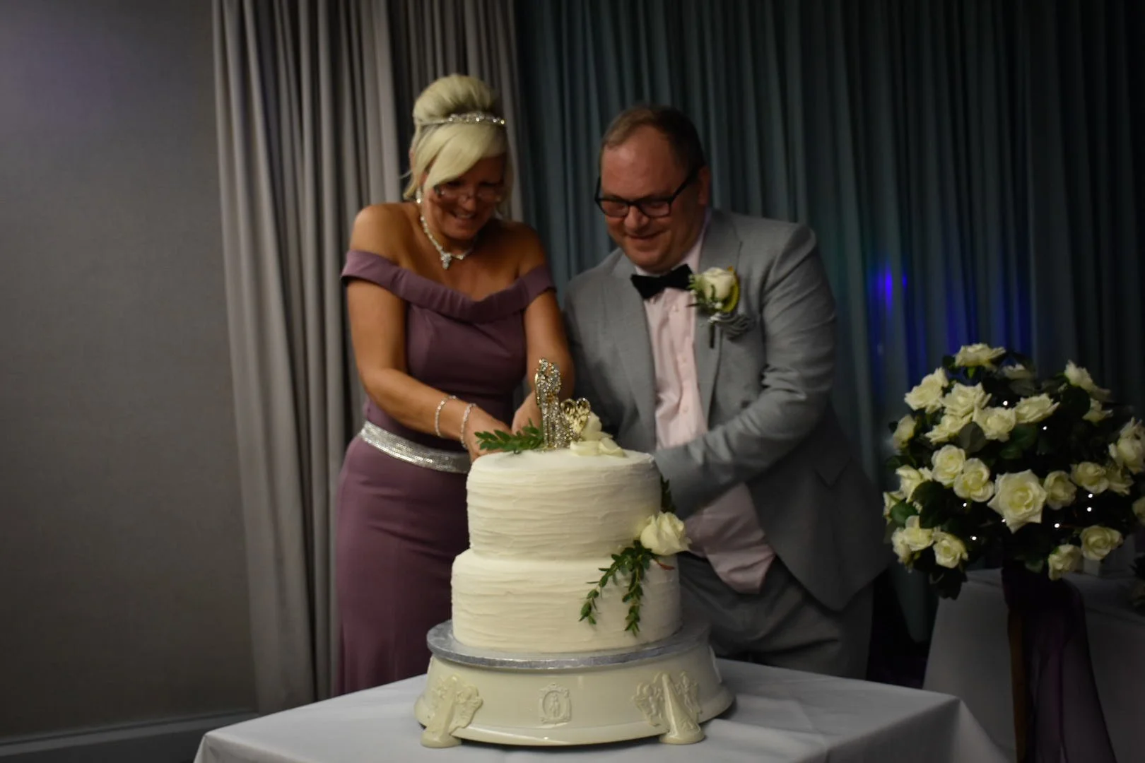 A woman and a man, dressed in formal wedding attire, are cutting a wedding cake together at a reception.