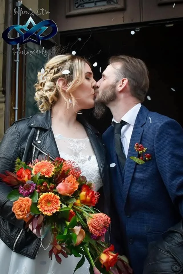 A couple shares a kiss on their wedding day, with the bride holding a large bouquet of colorful flowers and wearing a lace dress and leather jacket, while the groom wears a blue suit and tie, with confetti falling around them.