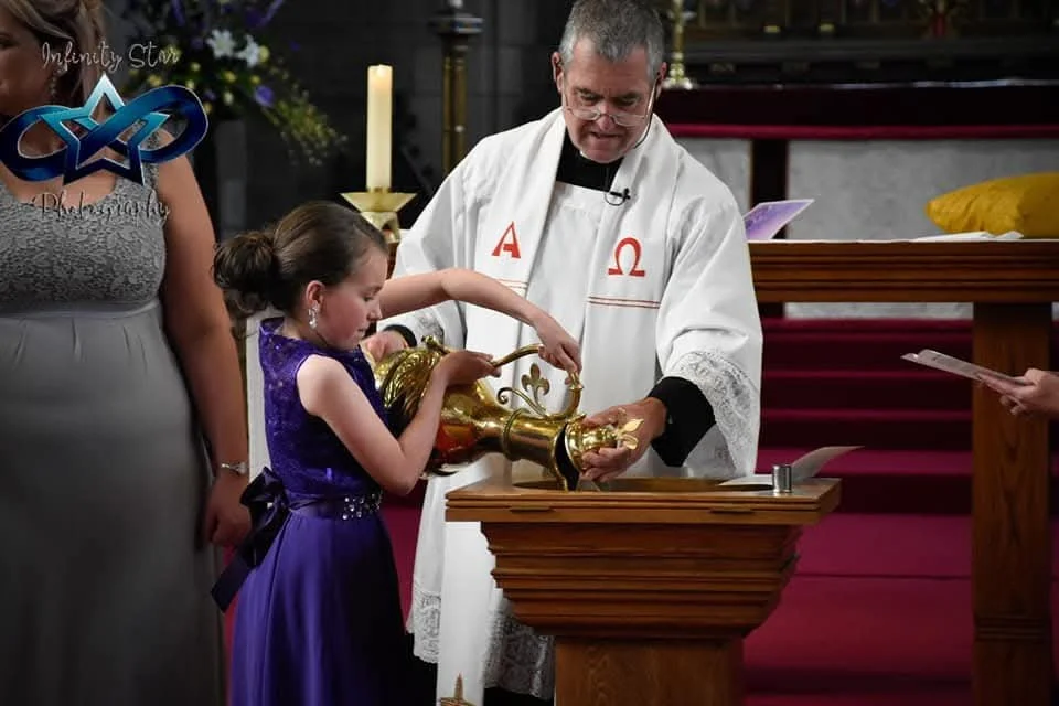 A priest in a white robe with Greek letters Omega and Alpha is pouring water from a gold vessel over a young girl during a religious ceremony, with a woman in a gray dress standing nearby and another person holding a paper.