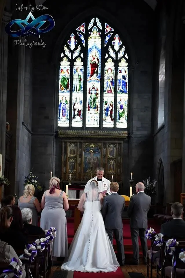 A wedding ceremony inside a church with a bride, groom, and officiant standing at the altar. The bride is wearing a white gown and veil, and the groom and guests are dressed formally. A large stained glass window with religious imagery is behind the altar, casting colorful light into the church.