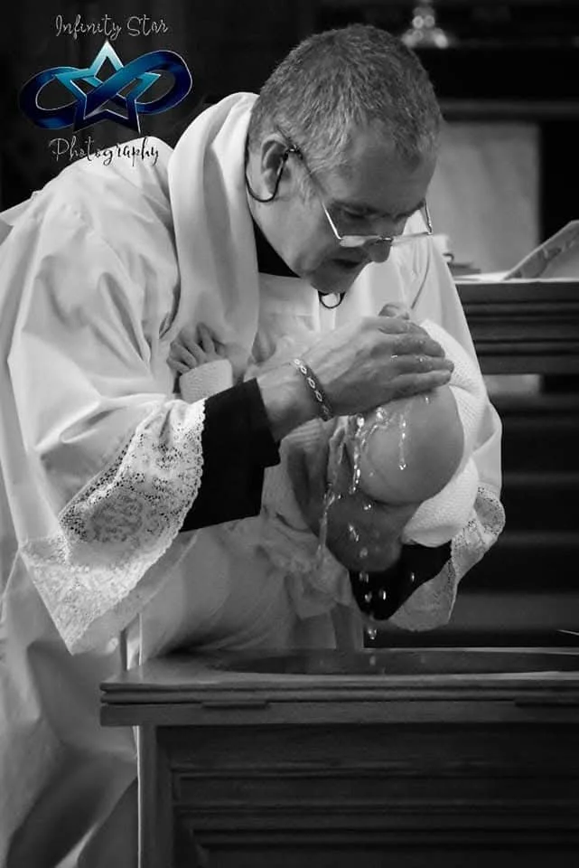 A man in religious attire baptizing a baby in a church setting, water pouring over the baby's head.