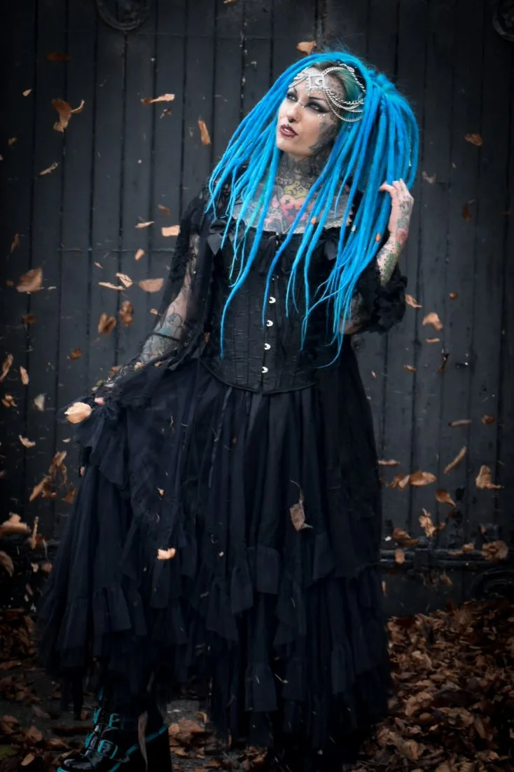 A woman with long blue dreadlocks, tattoos, and gothic makeup, wearing a black corset and layered black skirt, standing against a black wooden fence with falling leaves around her.