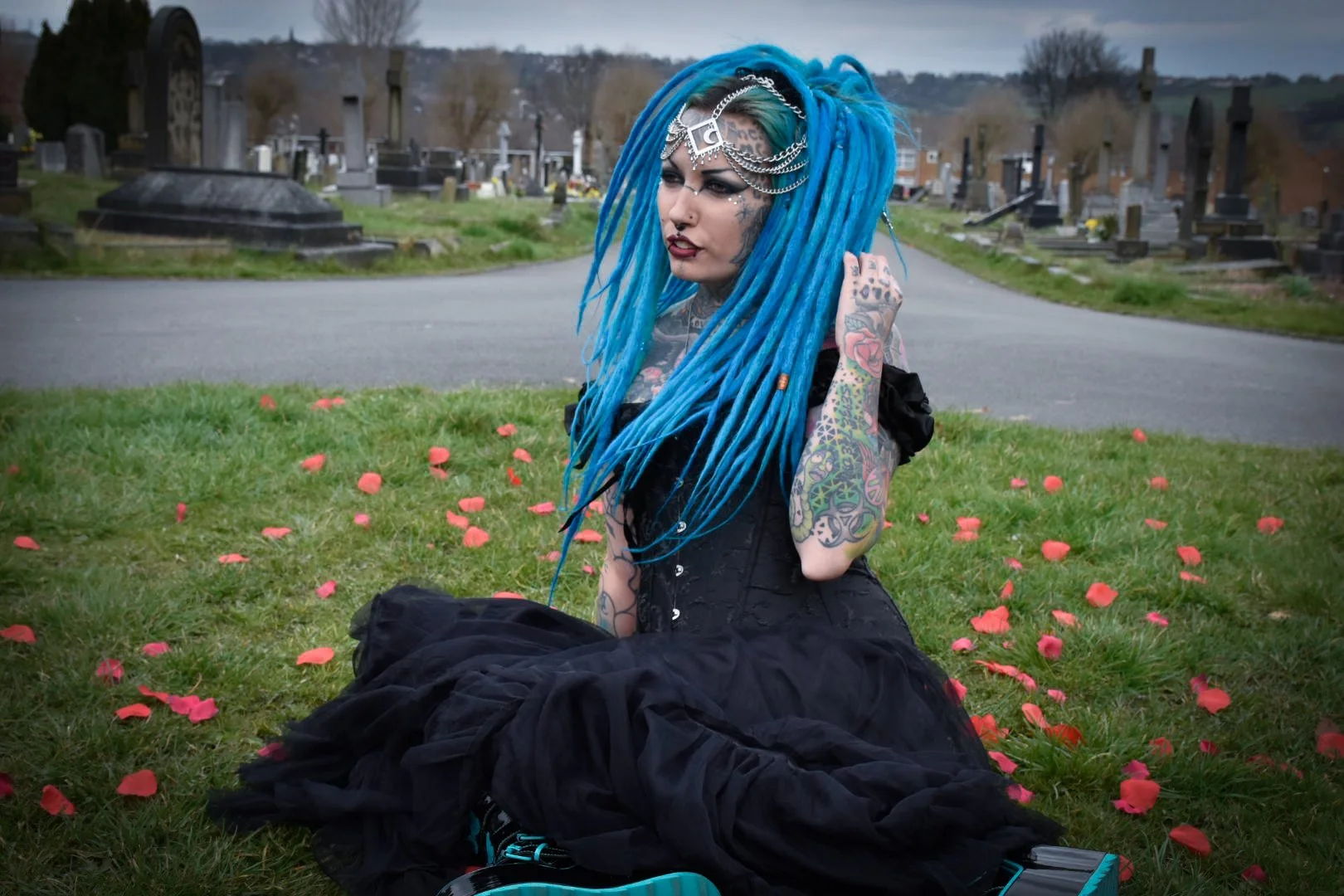 A woman with long, vibrant blue dreadlocks and tattoos on her arms sits on green grass scattered with pink flower petals in a cemetery. She wears black clothing and a silver chain headpiece, with dark lipstick and makeup, and appears to be lost in thought.