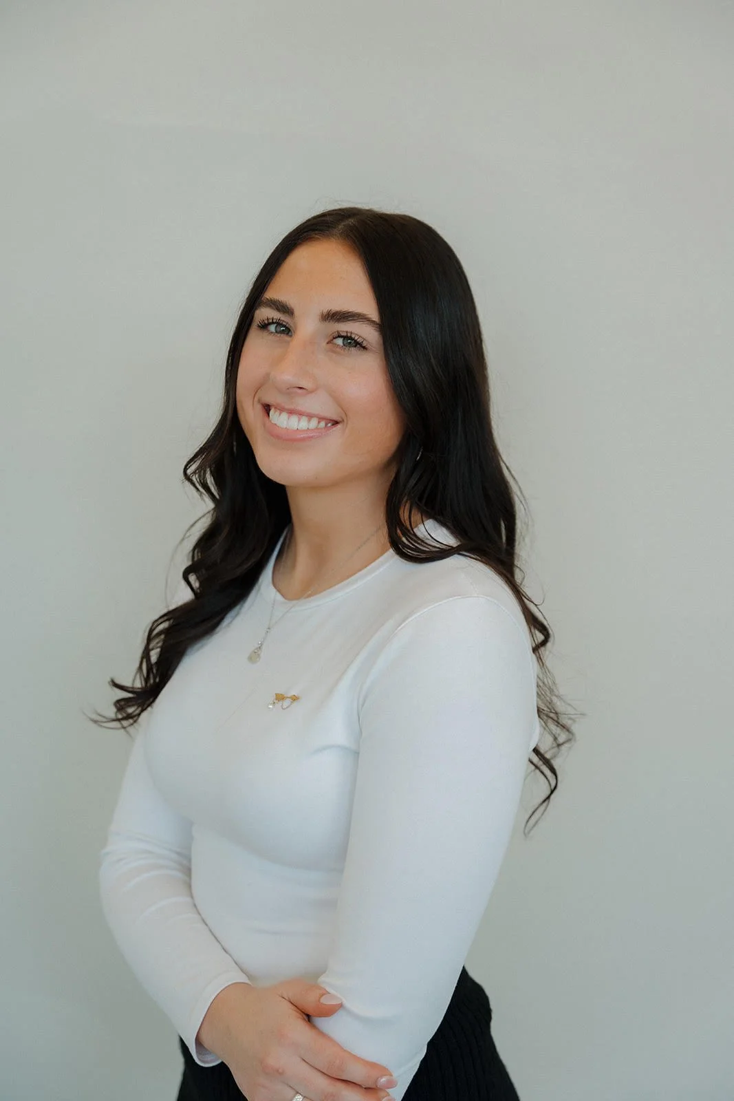 A young woman with long dark hair smiling in front of a plain light-colored wall, wearing a white long-sleeve shirt and a gold necklace with a small pendant.