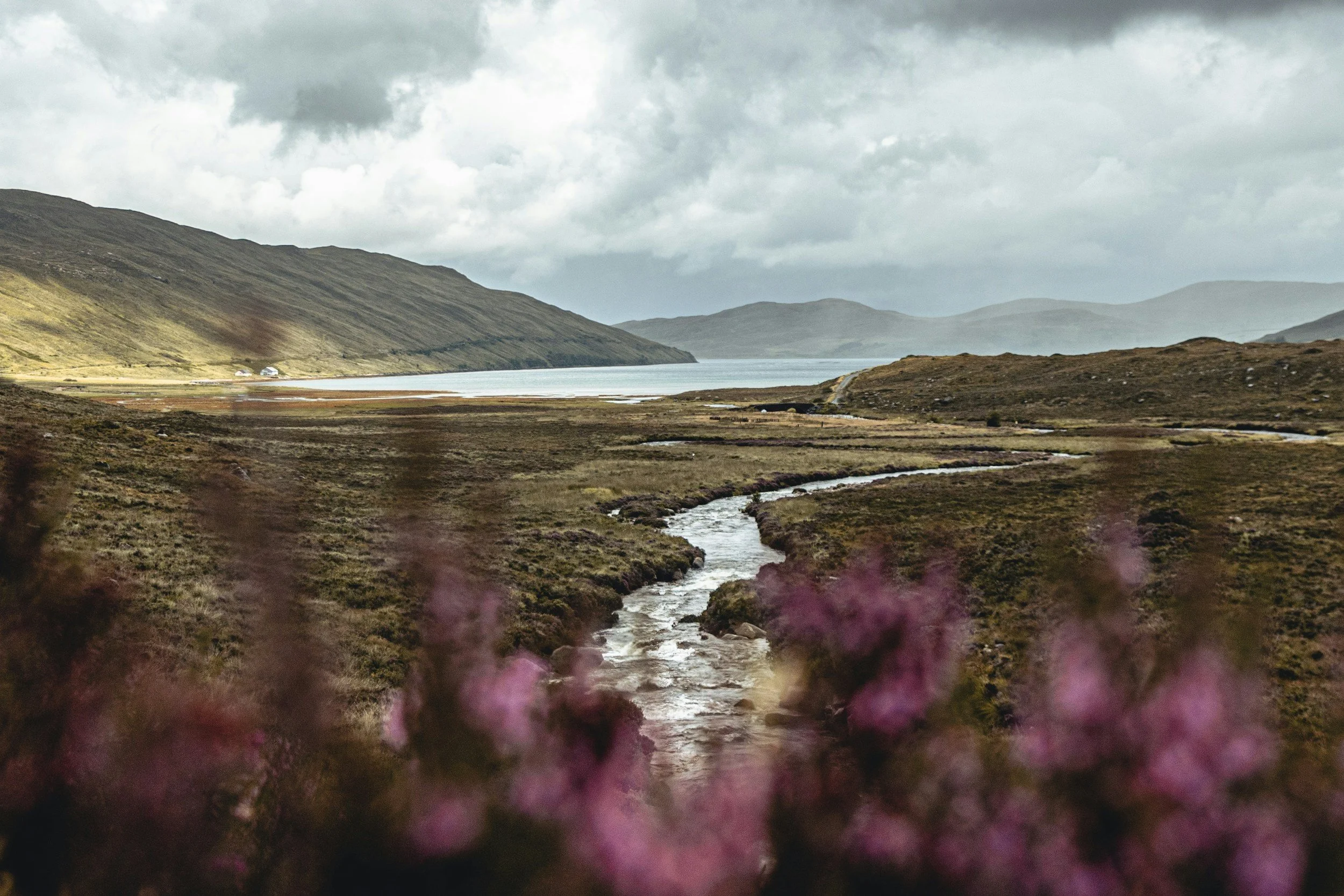 A mountainous landscape with a river flowing through a valley, cloudy sky, and some buildings in the distance.