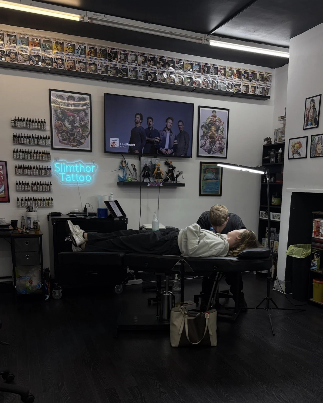Tattoo studio with two people, one lying on a table and the other attending, surrounded by artwork, figurines, and tattoo supplies.