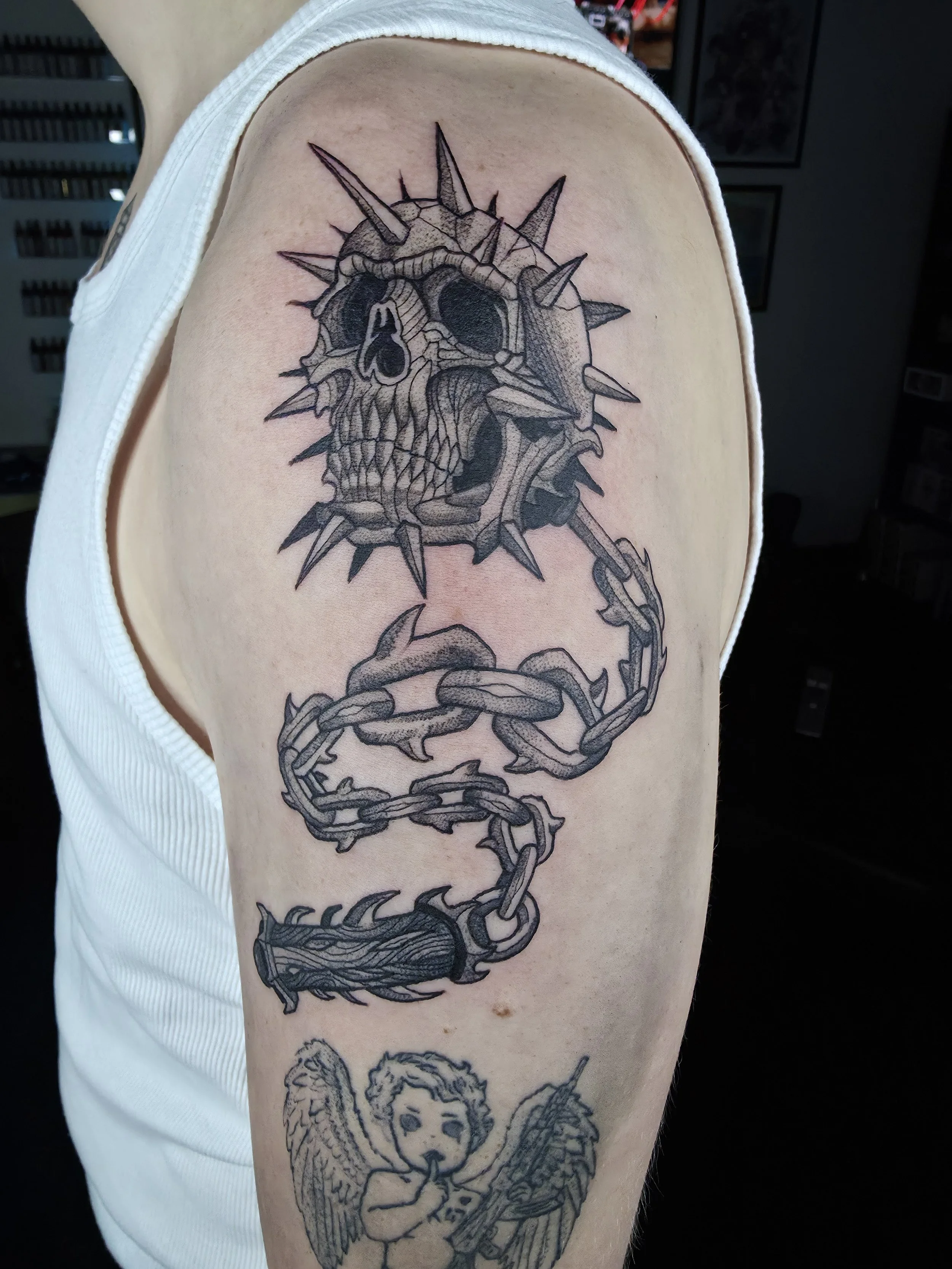 Black and gray tattoo on upper arm featuring a skull with spikes, chains, and a winged figure.