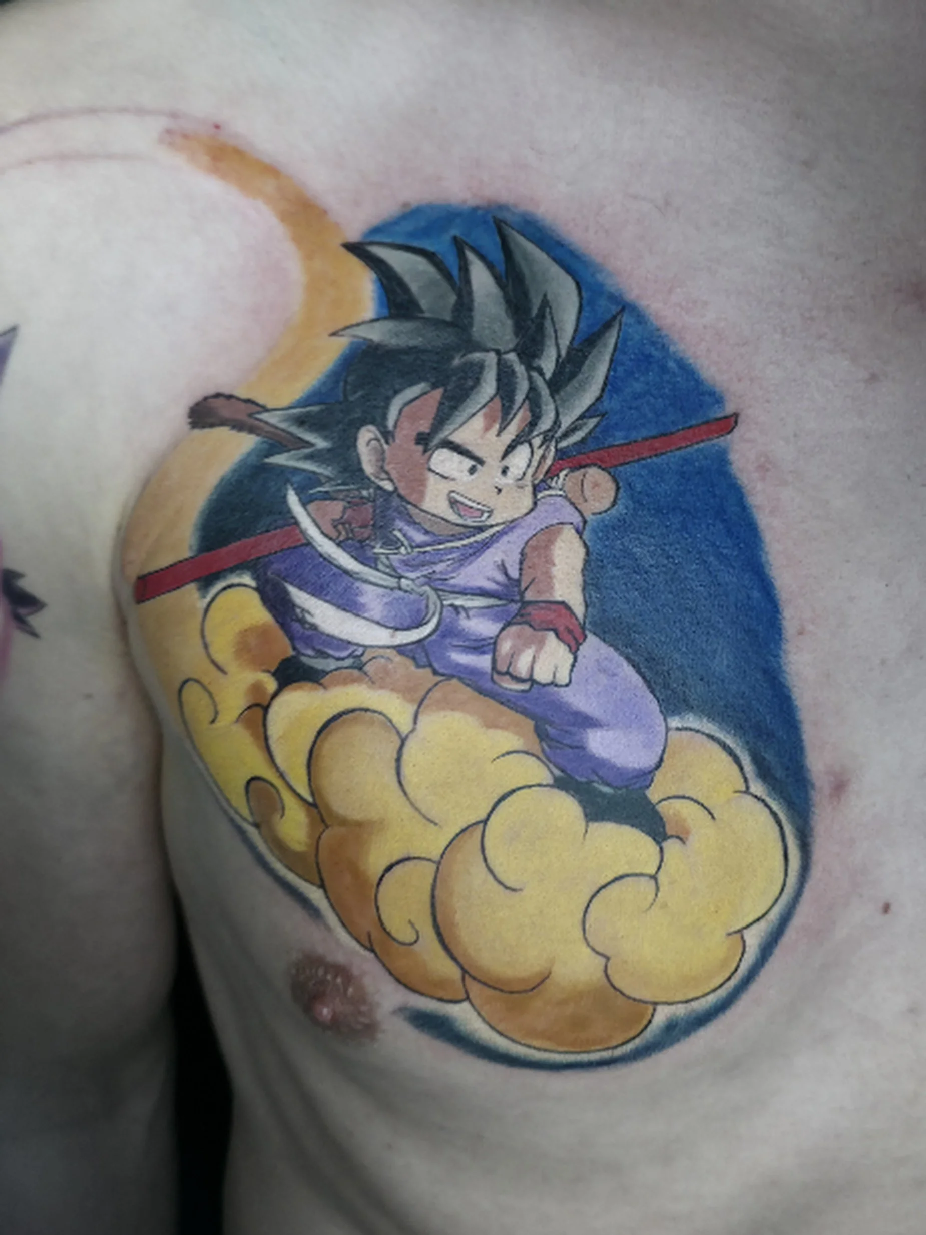 A tattoo of Goku from Dragon Ball z, mid-air with a blue background, yellow clouds beneath him, and a red power pole in his hand.