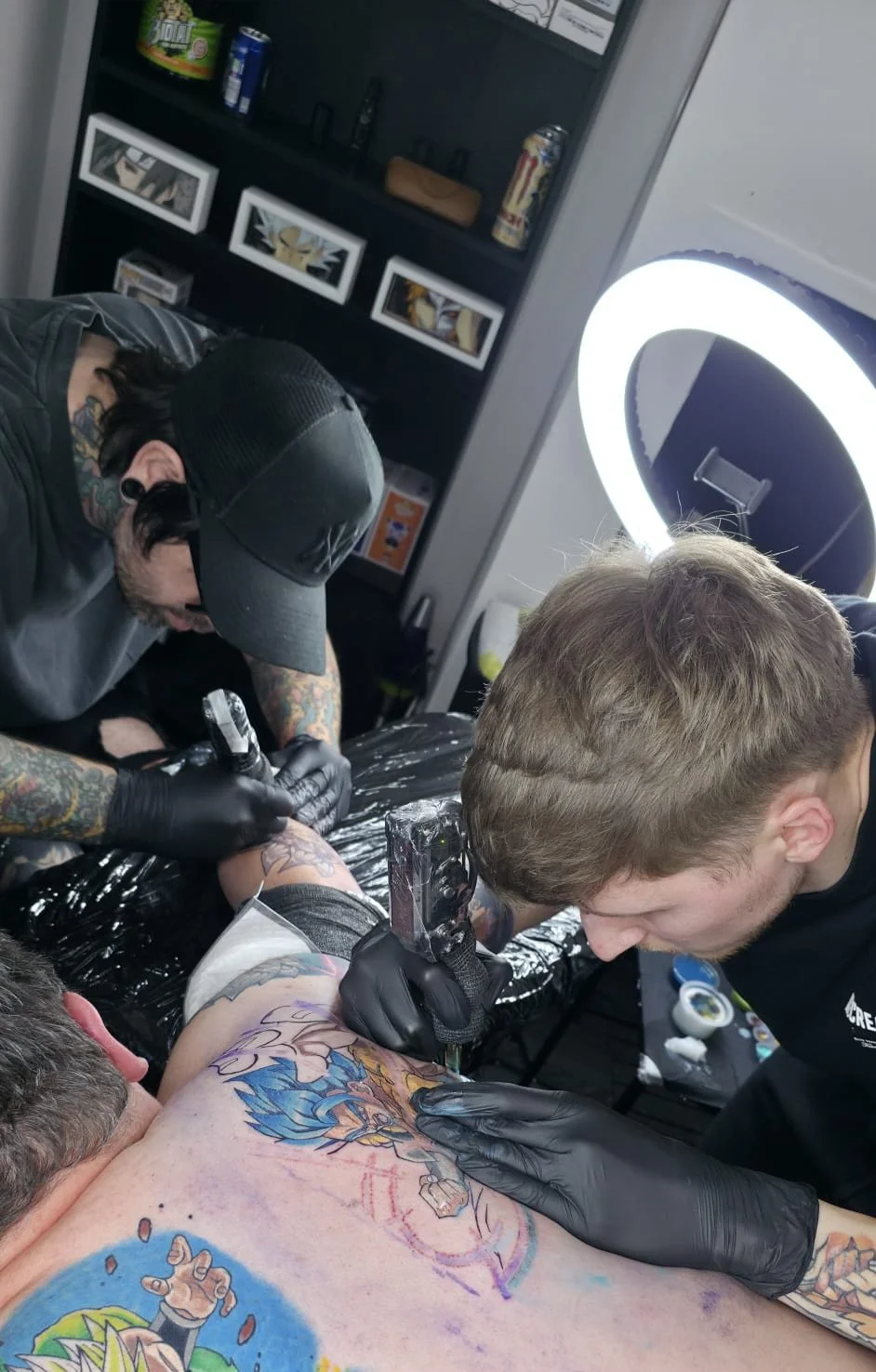 A person getting a colorful tattoo on their arm at a tattoo studio, with a tattoo artist and an assistant working on the tattoo while tattooing equipment is visible.