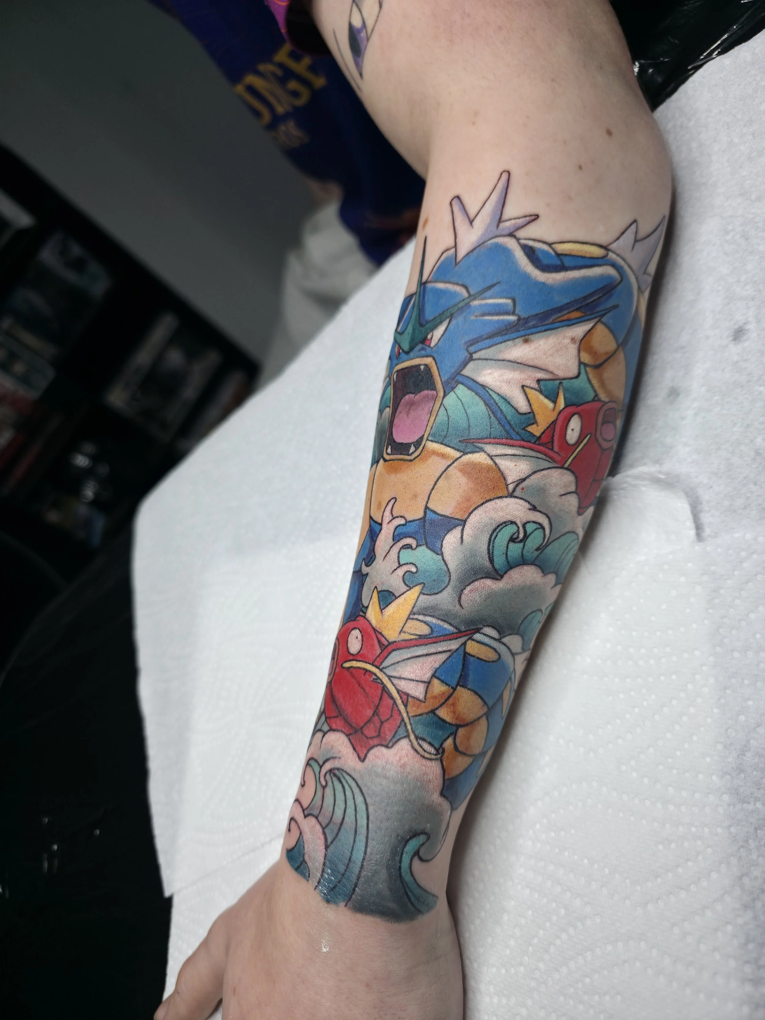 A colorful tattoo of three Pokémon characters on a person's arm. The Pokémon include Garchomp, Charizard, and Blastoise, depicted in a vibrant, cartoon-style design.