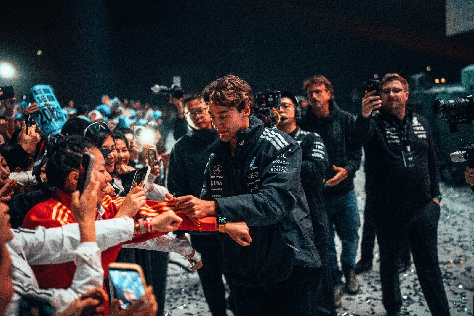 A race car driver in black team kit signing an autograph for young fans at night, surrounded by photographers and fans taking pictures.